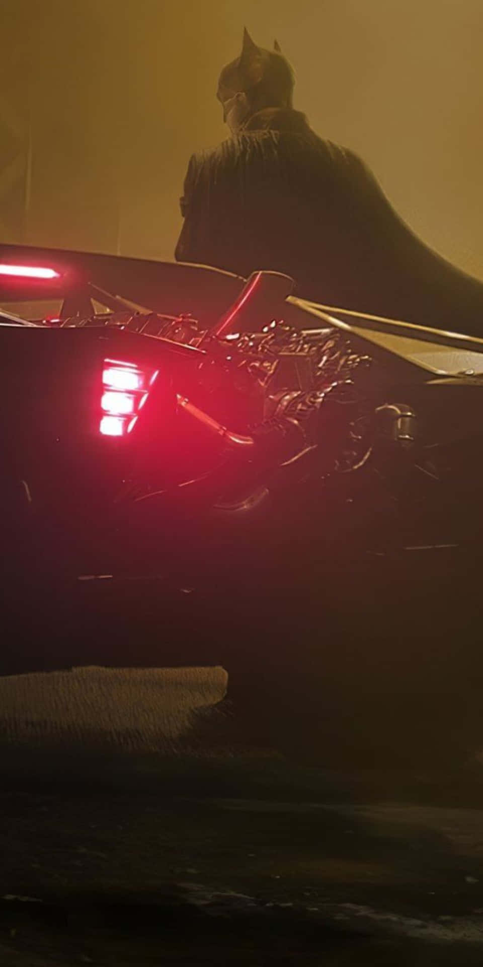 蝙蝠俠站在他的蝙蝠車後方，車燈發出紅光，周圍環境黑暗且煙霧瀰漫。