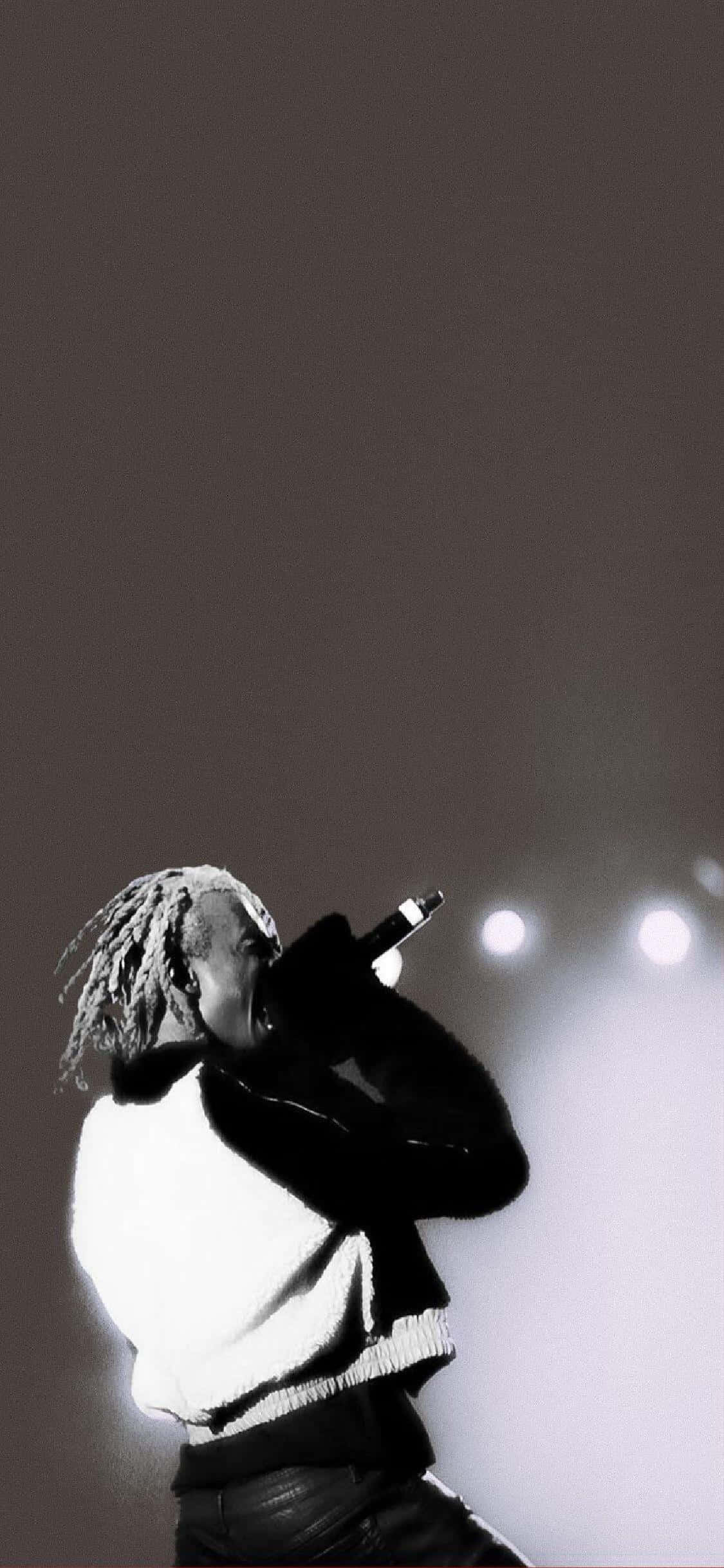 今天就入手 Playboi Carti 限量版 iPhone！ 壁紙 — 一位留著辮子的音樂家在明亮的舞台燈光下對著麥克風唱歌，這是一張黑白表演照。
