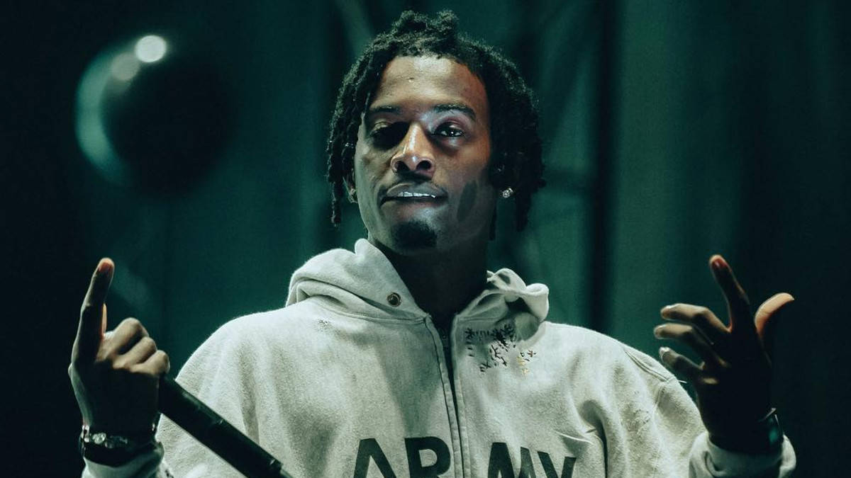 Playboi Carti PFP 穿連帽衫 壁紙 — 一名留著辮子頭、身穿白色連帽衫的男子，在昏暗的綠色舞台燈光下，一邊拿著麥克風一邊做手勢。