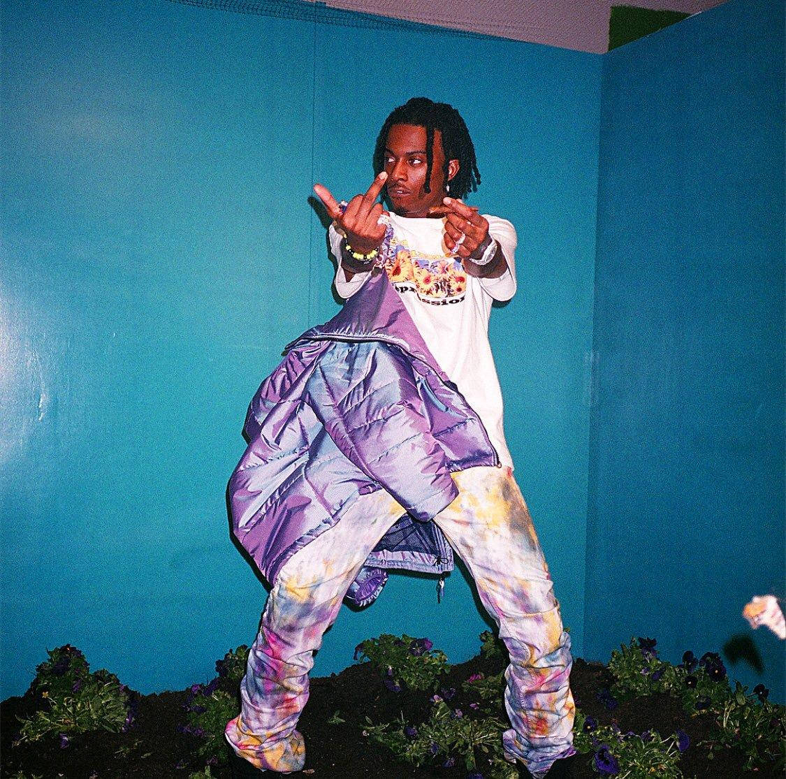 Playboi Carti PFP 紫色版 壁紙 — 一個人穿著鮮豔的紮染褲和紫色羽絨外套，在腳邊有紫色花朵的青色牆前擺姿勢。