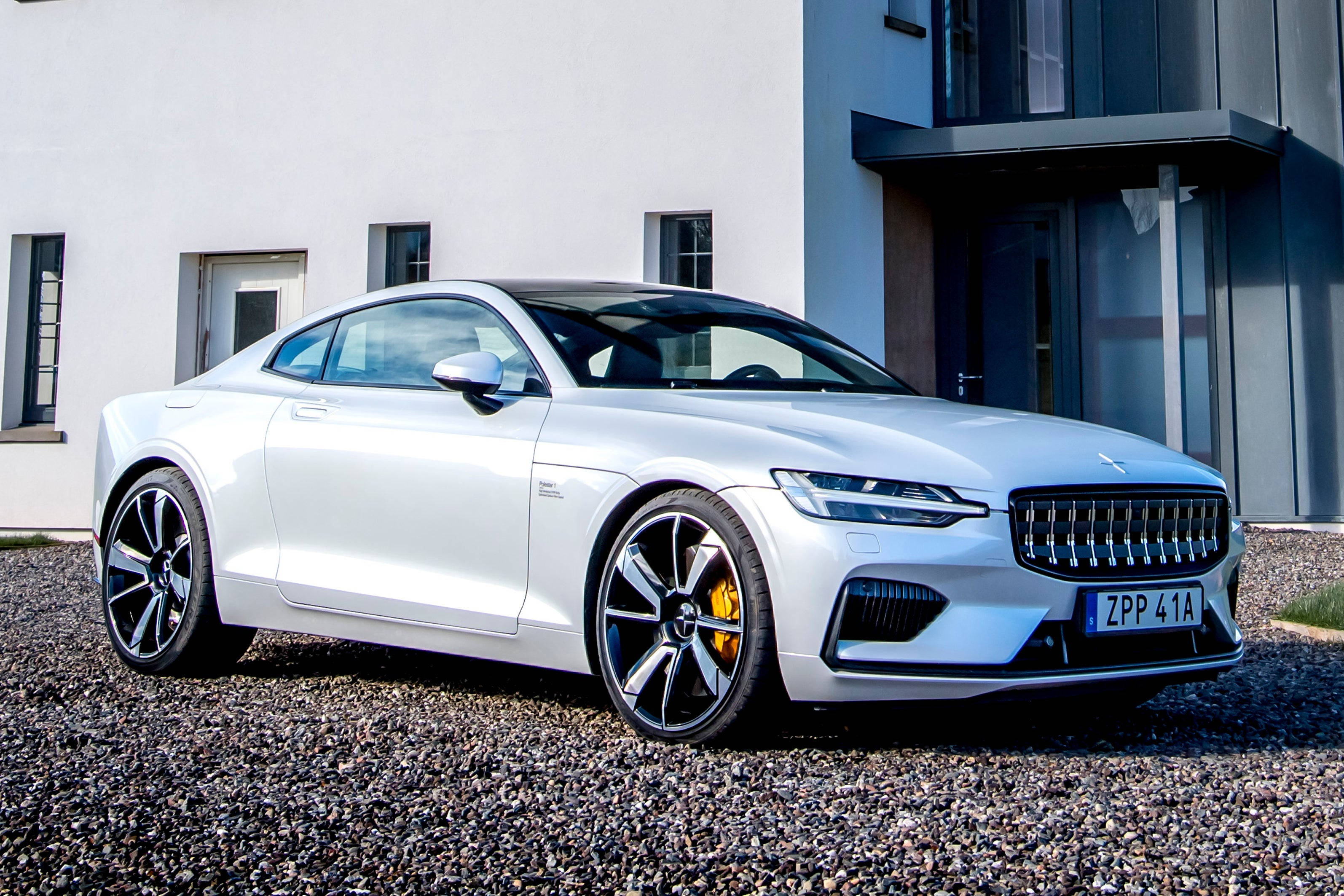 Polestar 1豪華車 白色版 壁紙 — 一輛時尚的白色電動轎跑車，配備黑色輪圈和黃色制動卡鉗，停在現代建築前的礫石上。