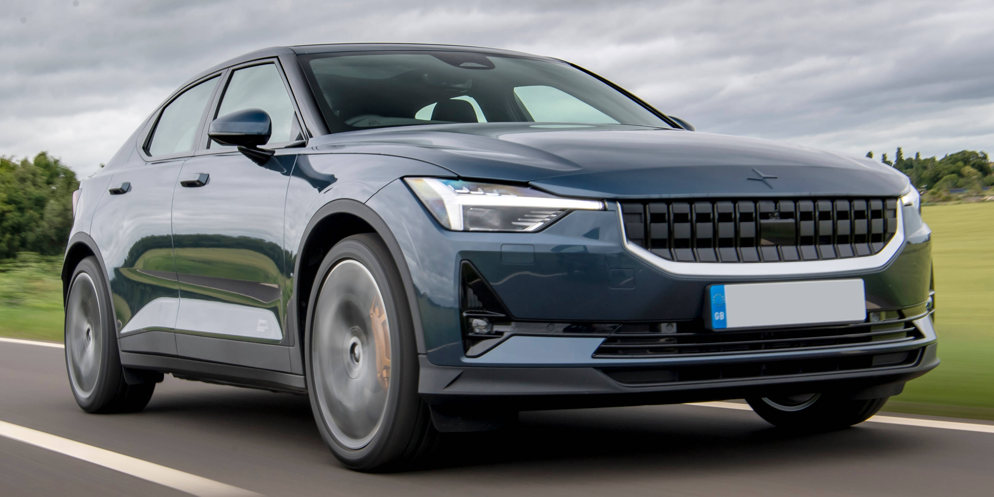 在晴朗日光下展現流線型的Polestar 2電動車 壁紙 — 一輛時尚的深藍色電動車在鄉間道路上疾馳，背景是模糊的綠色田野和陰天。
