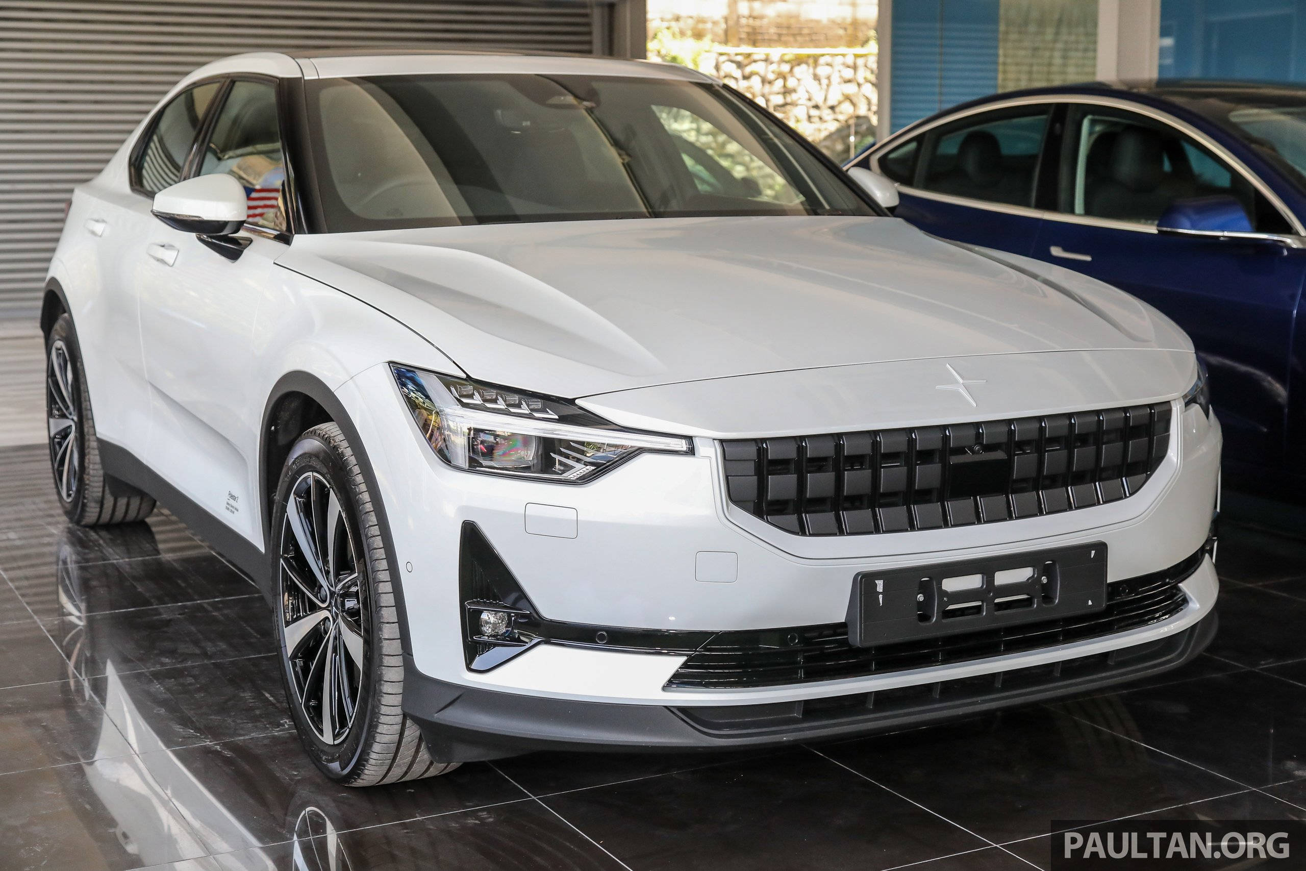 雪色 Polestar 2 壁紙 — 一輛時尚的白色Polestar 2電動車在展廳燈光下閃閃發光，後面可見一輛深藍色轎車。