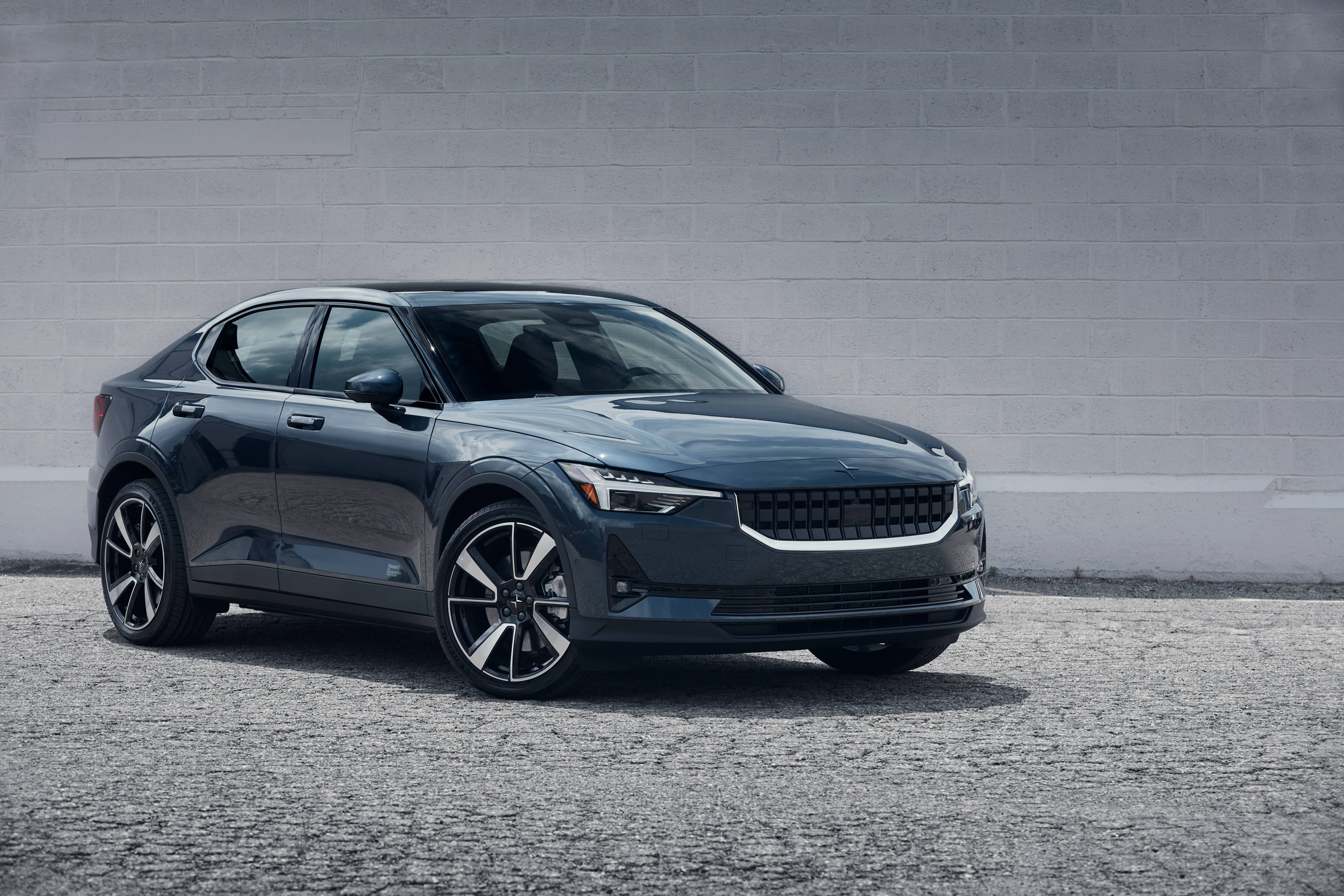 Polestar 2 深灰色版 壁紙 — 一輛時尚的深灰色現代轎車在明亮的日光下停泊在碎石上，背景是樸素的灰色磚牆。