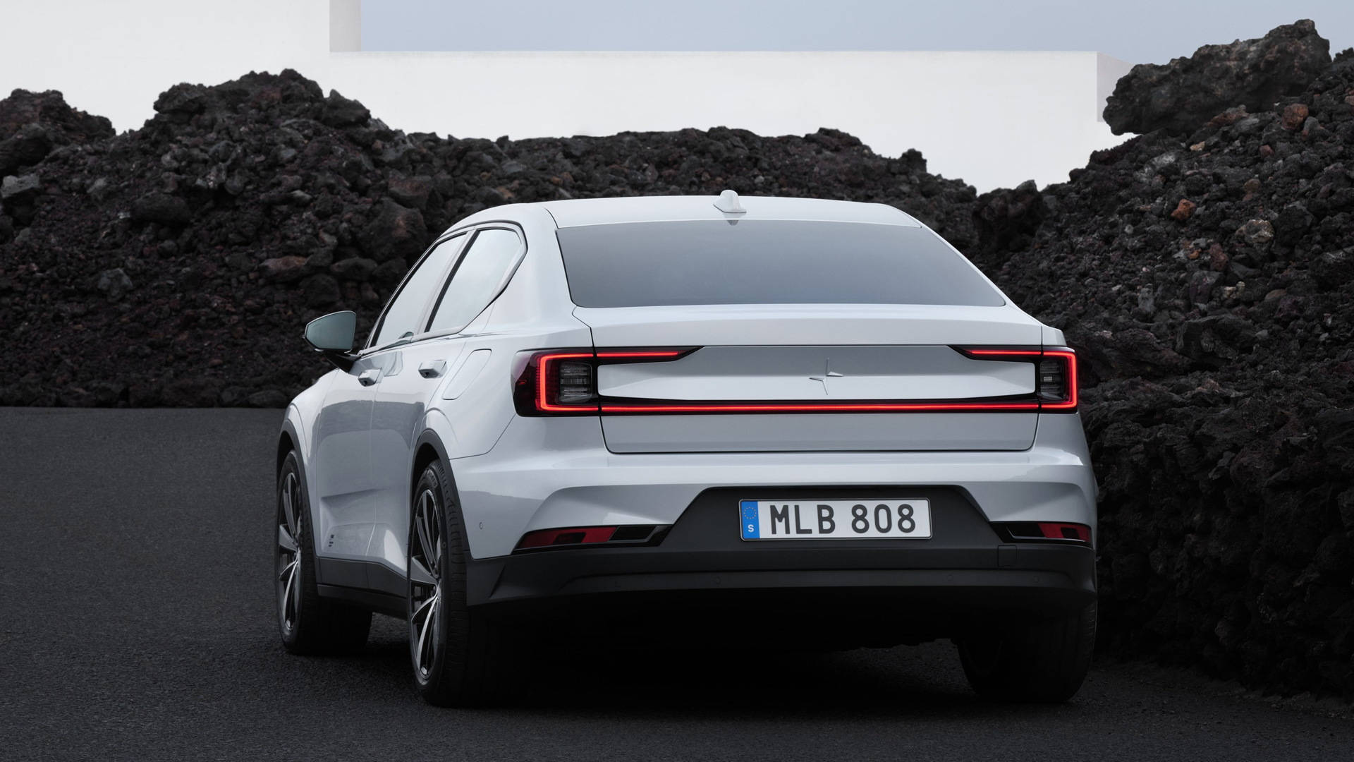 Polestar 2 行駛於岩石路 壁紙 — 一輛時尚的白色電動車，帶有發光的紅色尾燈，停在瀝青路面上，旁邊是深色的火山岩層。