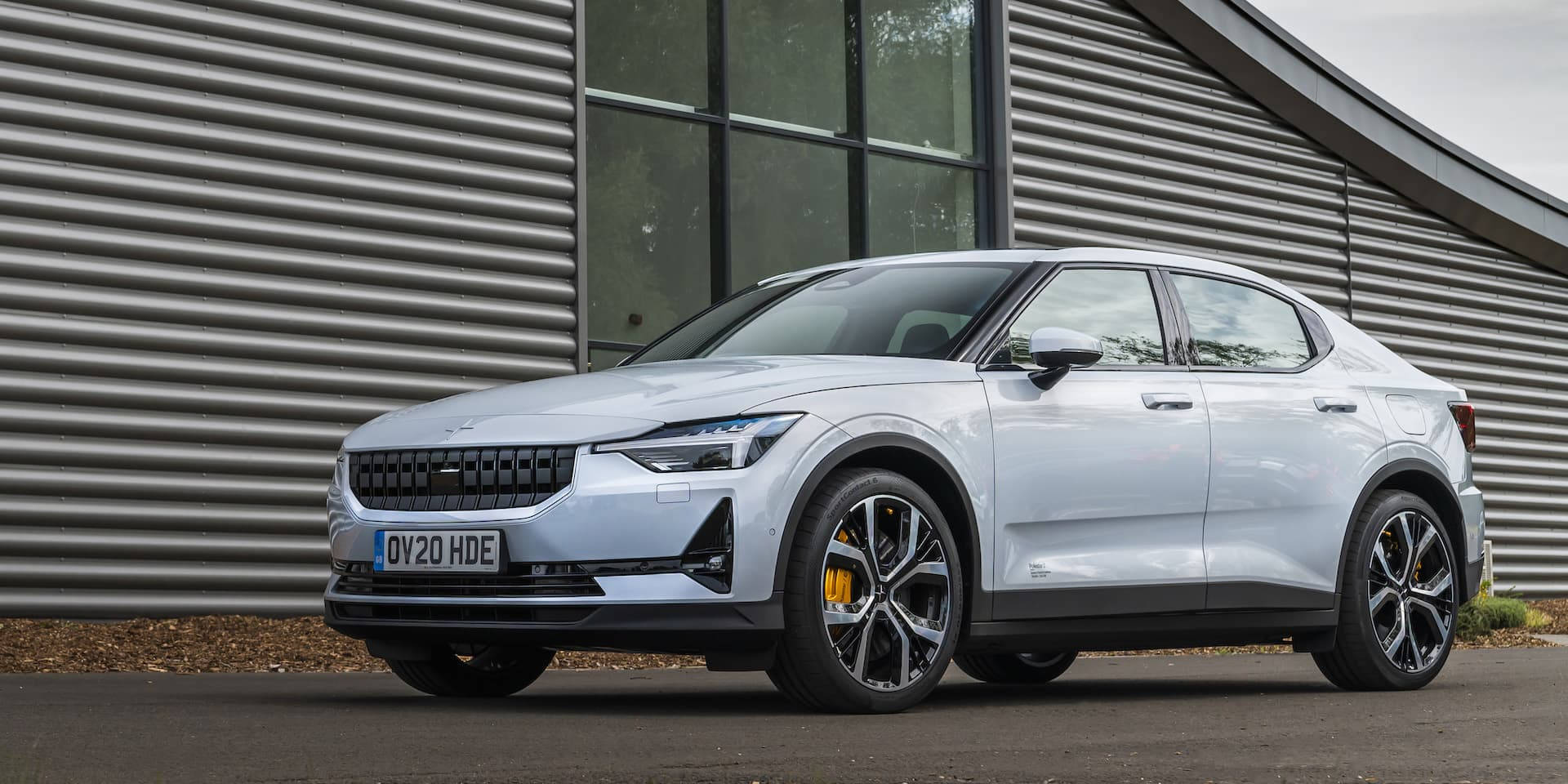 Polestar 2停泊現代住宅旁 壁紙 — 一輛時尚的銀色電動車，帶有黃色制動卡鉗，停在現代波紋金屬建築前。