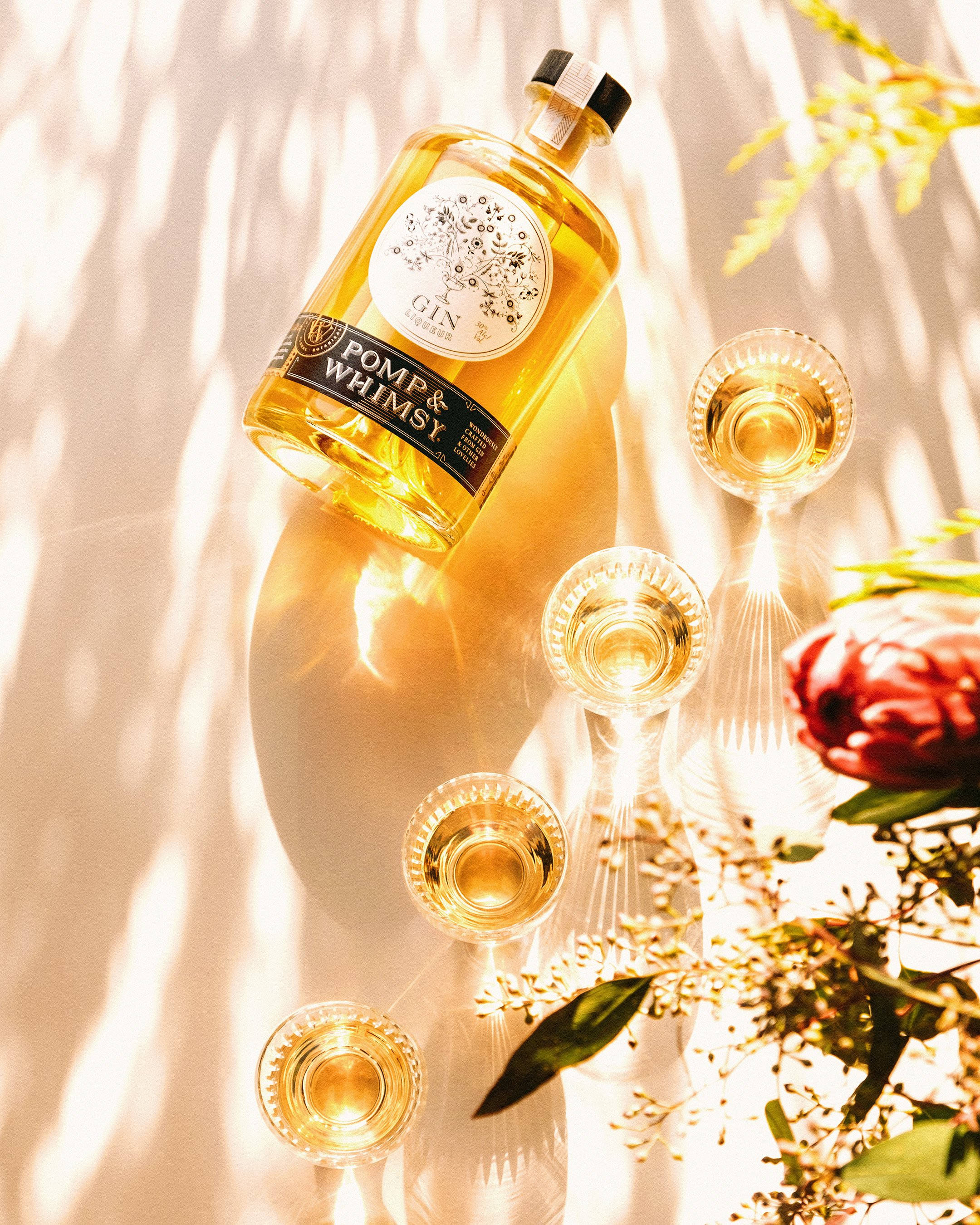 Pomp & Whimsy 飲料 壁紙 — 金色氈酒瓶和酒杯在溫暖的陽光下透過樹葉照射，周圍環繞著鮮花和植物。