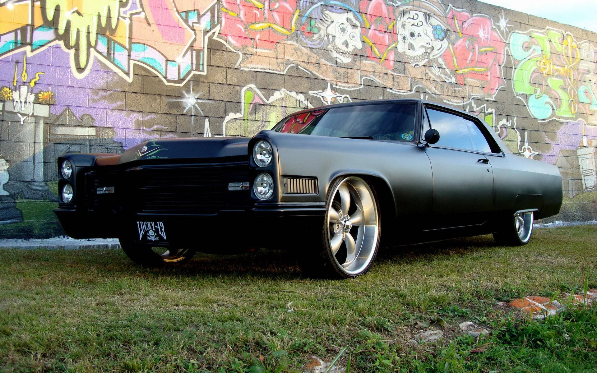 Pontiac GTO 磨砂黑 壁紙 — 一輛啞光黑色的經典肌肉車，配備定制輪轂，停在草地上，前面是一幅充滿活力的塗鴉牆。