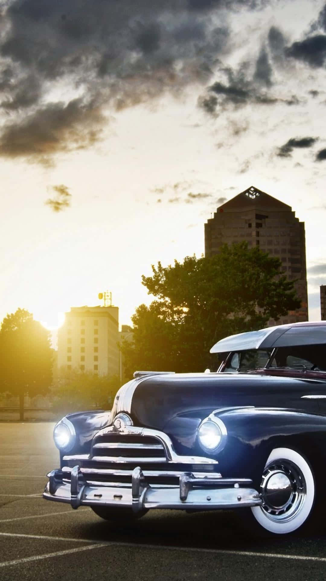 經典 Pontiac Streamliner 跑車 iPhone 壁紙 — 一輛經典藍色汽車，帶有鍍鉻細節和白色車壁，停放在城市停車場，背景是日落時分的戲劇性雲彩。