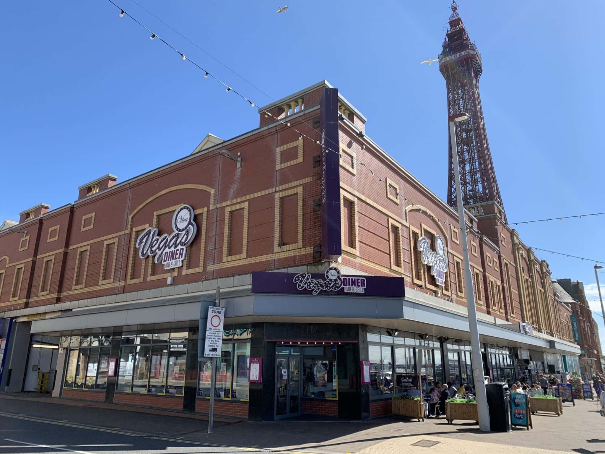 從城市拍攝的黑pool塔照片 壁紙 — 晴朗嘅日子，喺紅磚 Vegas Diner，後面係 Blackpool Tower 喺藍天白雲下高聳。