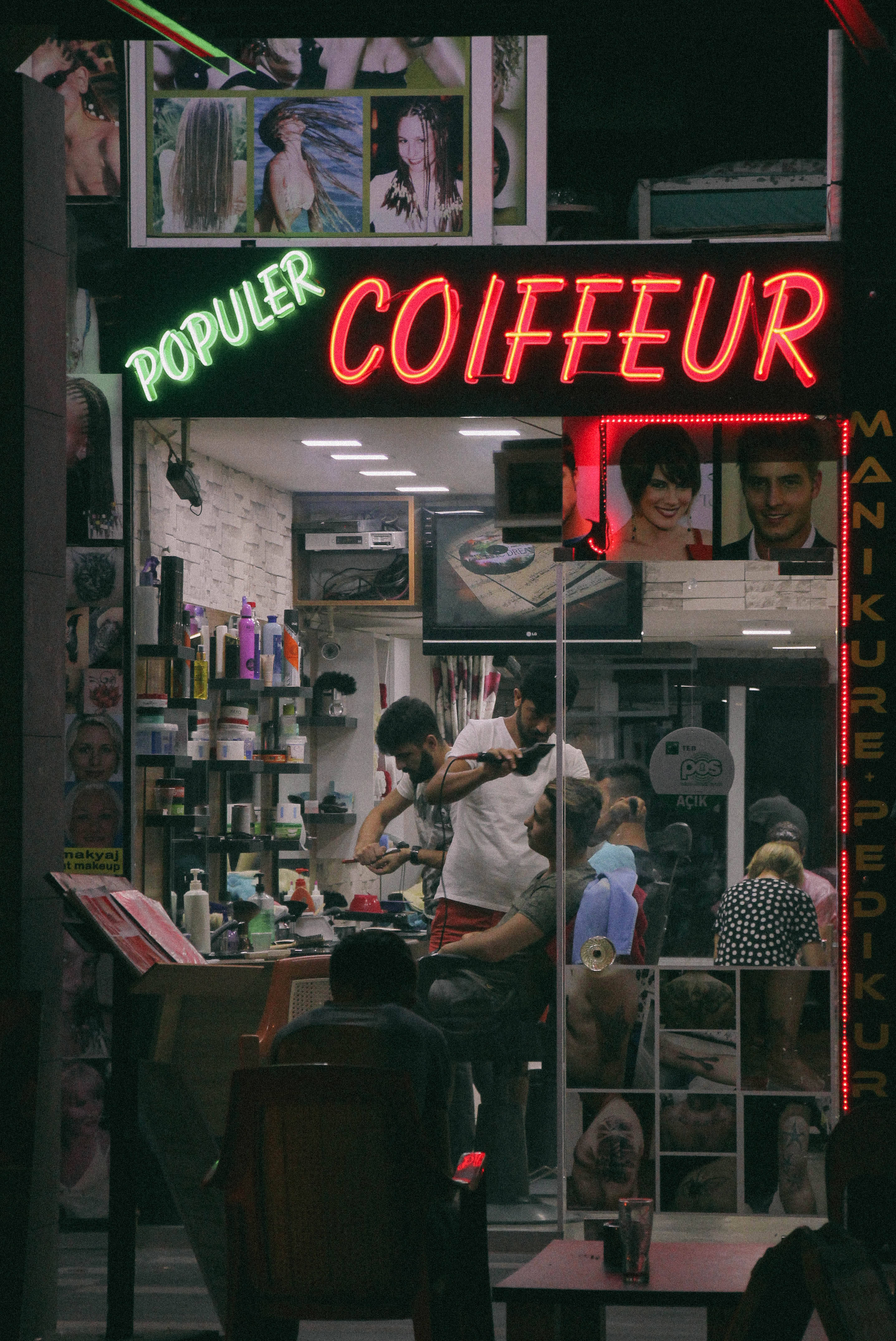 Populer Coiffeur髮廊 壁紙 — 夜晚一間充滿活力的霓虹燈髮廊，繁忙的市區店面內，造型師正在為顧客服務。