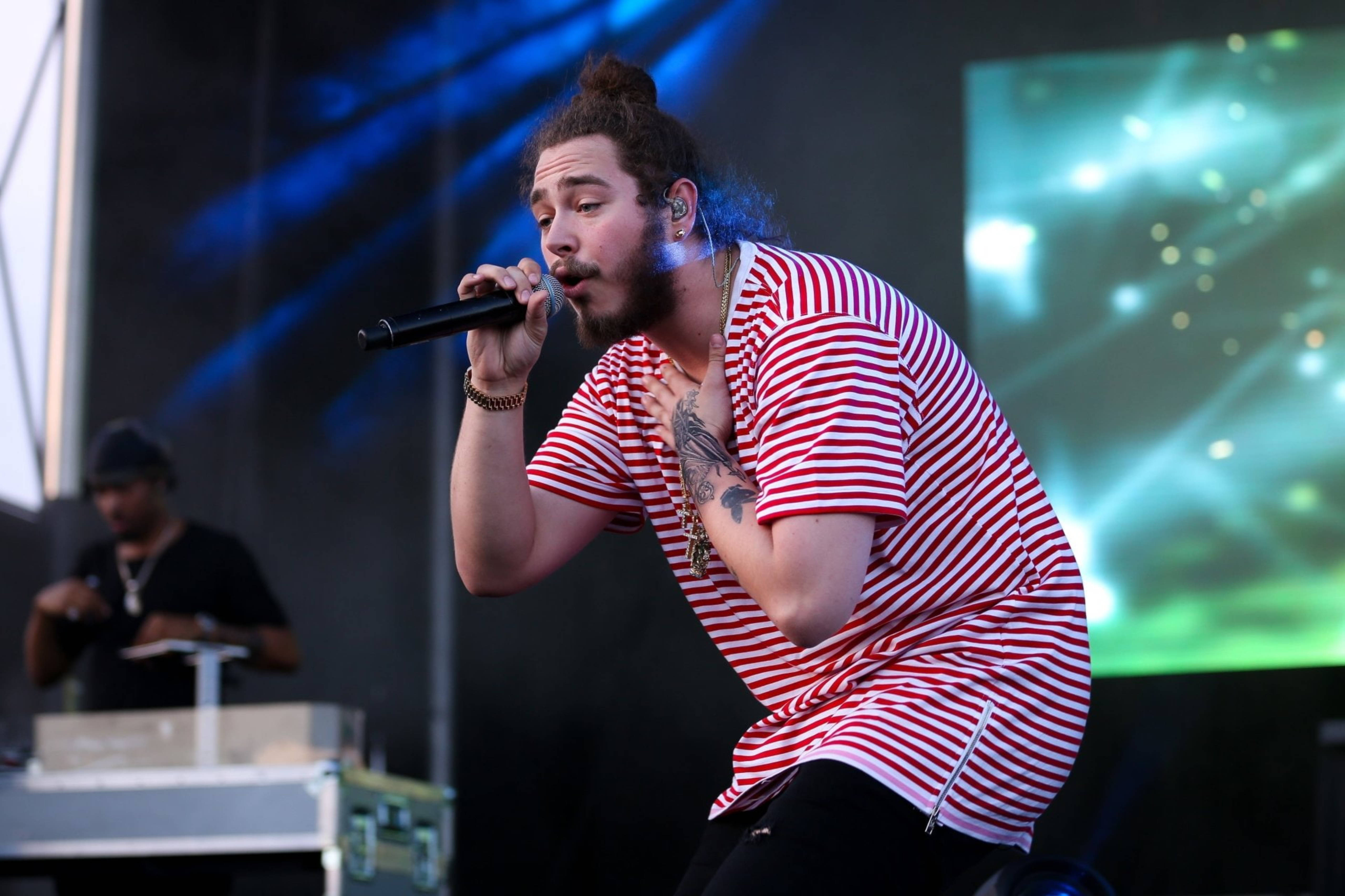 Post Malone 2016達拉斯現場演出 壁紙 — 一位留鬍子、束髮髻的男子在燈光璀璨的舞台上，對著咪高峰熱情獻唱，舞台燈光呈藍色和綠色。