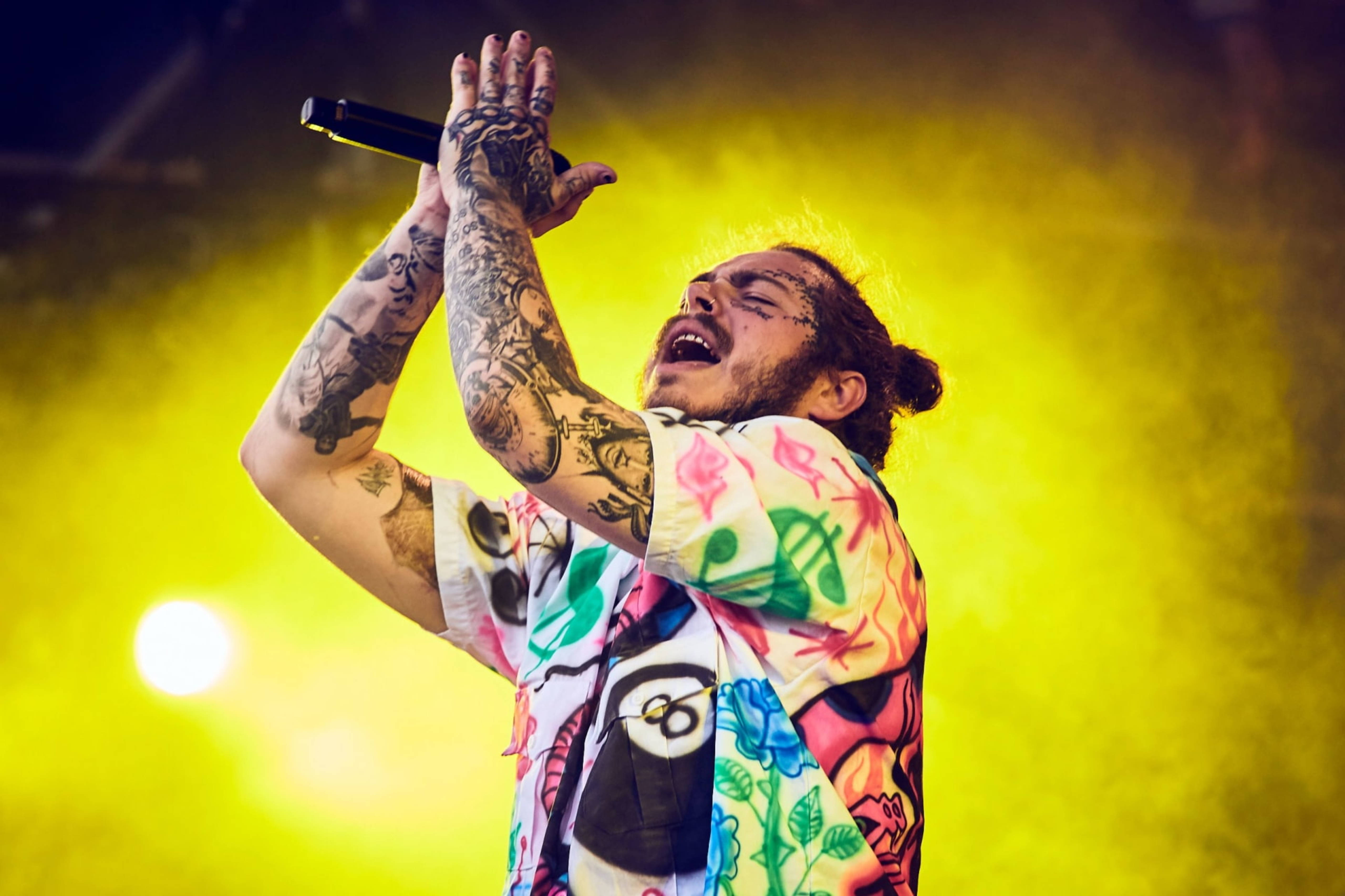 Post Malone 2018芝加哥Lollapalooza音樂節 壁紙 — 一位紋身表演者閉著眼睛，在強烈的黃色舞台燈光下熱情地對著麥克風唱歌。