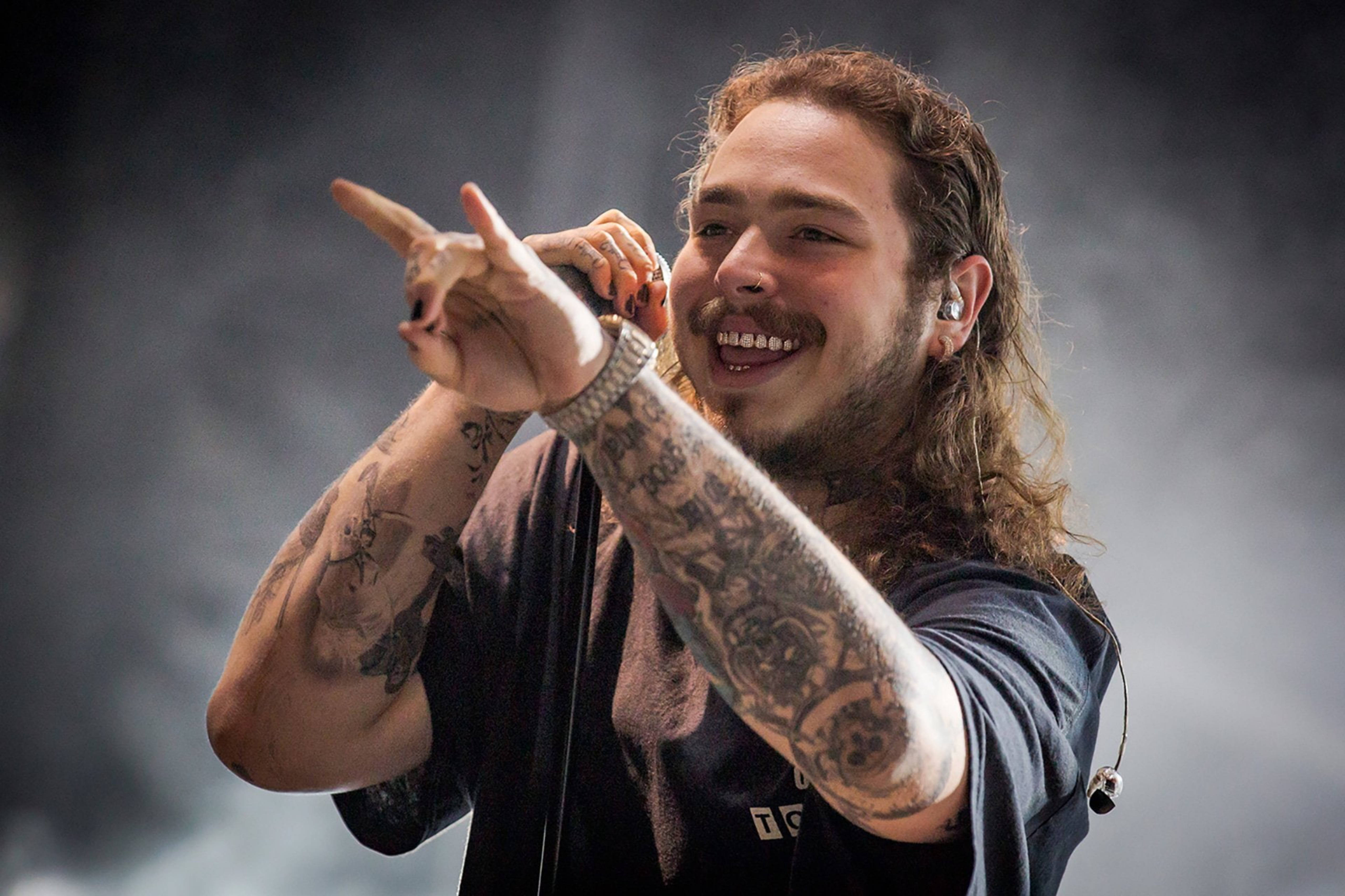 Post Malone 2018羅馬Rock In Roma演唱會 壁紙 — 一位紋身長髮音樂家在煙霧彌漫的舞台上微笑並充滿活力地做手勢表演。