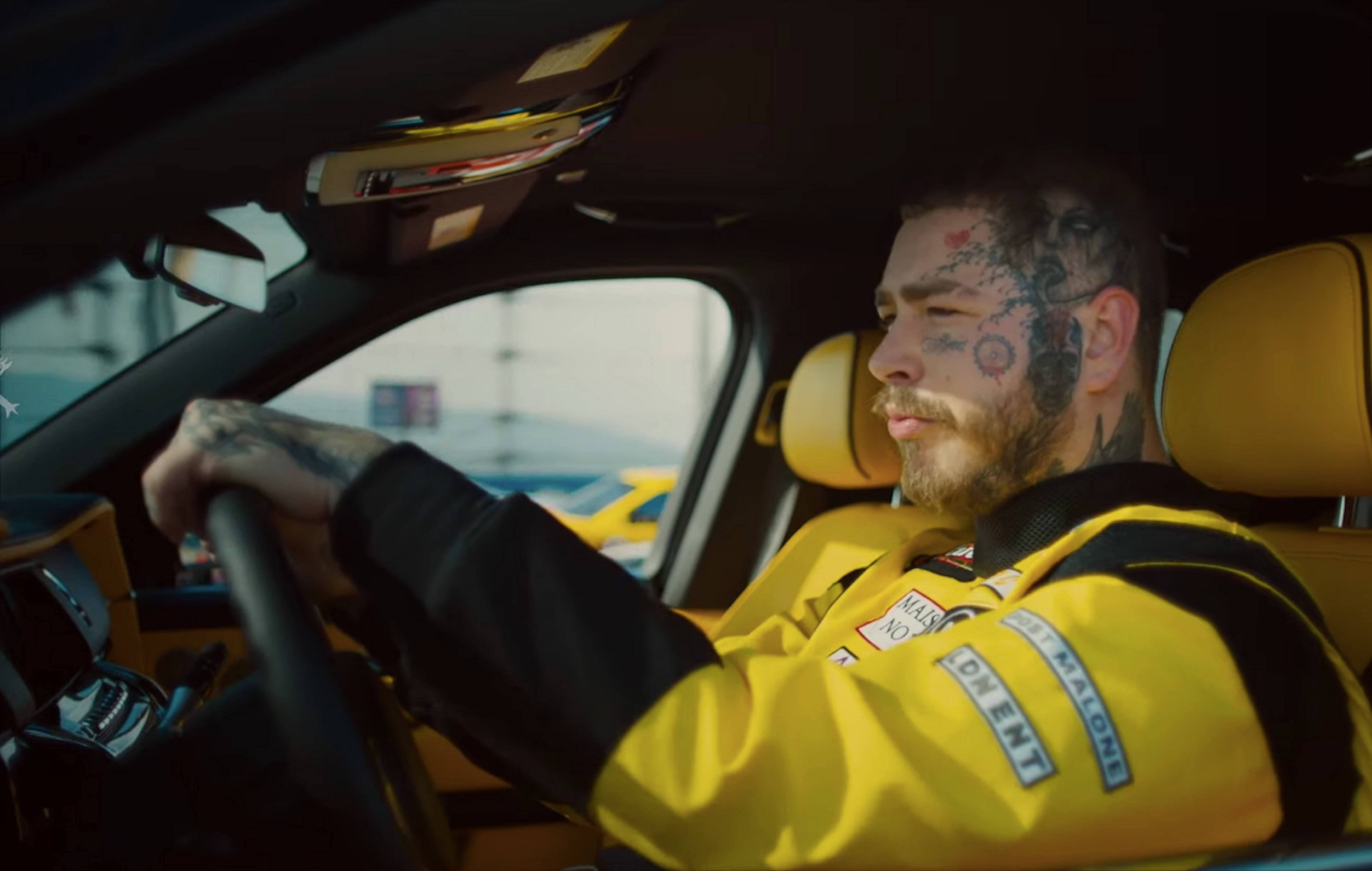 Post Malone《Motley Crew》音樂錄影帶場景 壁紙 — 一位紋身男子身穿黃色賽車服，專注地握著黃色賽車內的方向盤。