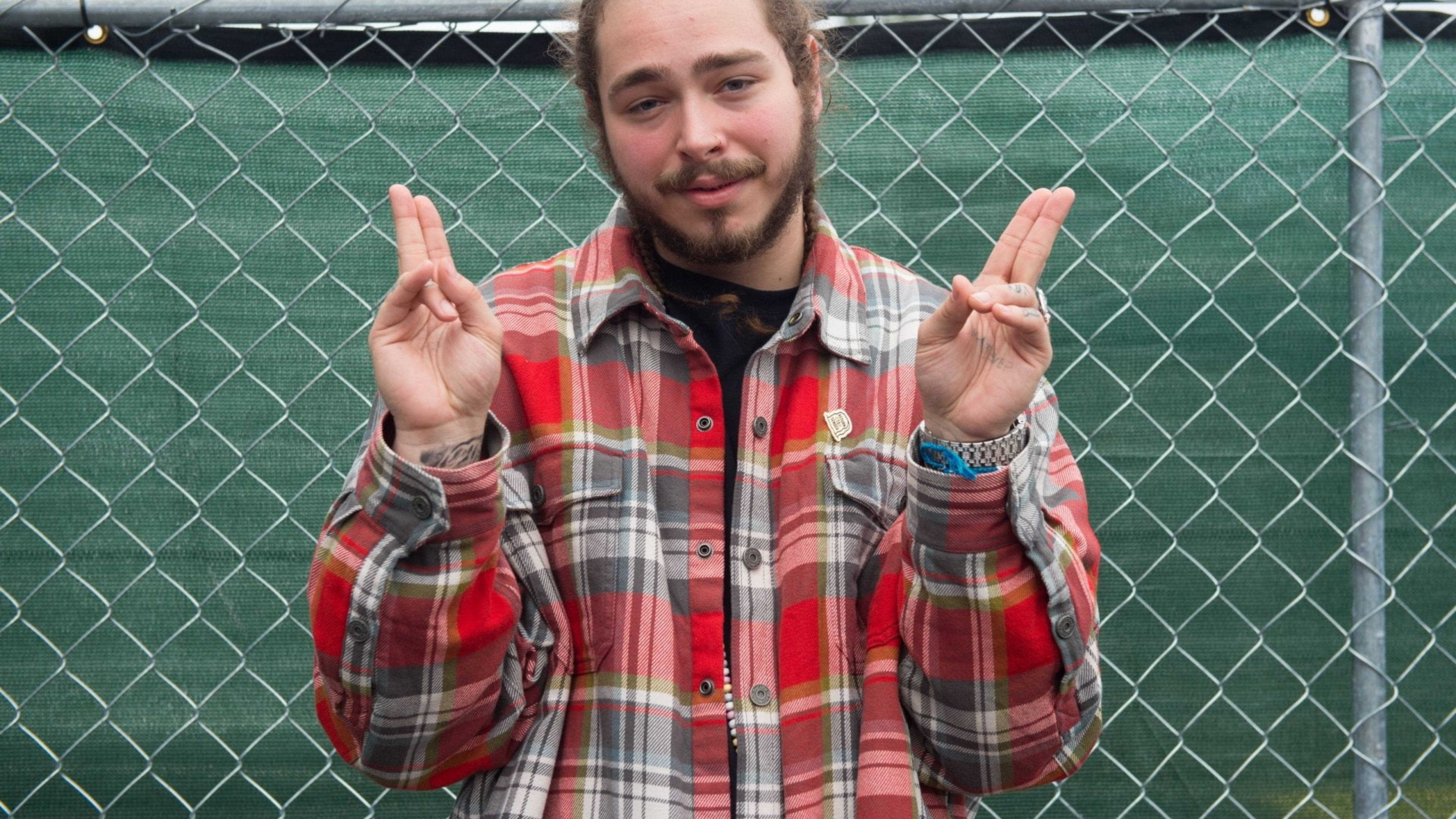 Post Malone The Meadows 音樂藝術節 2016 壁紙 — 一名留鬍鬚的年輕男子穿著格仔恤衫，在綠色鐵絲網前做出和平手勢。