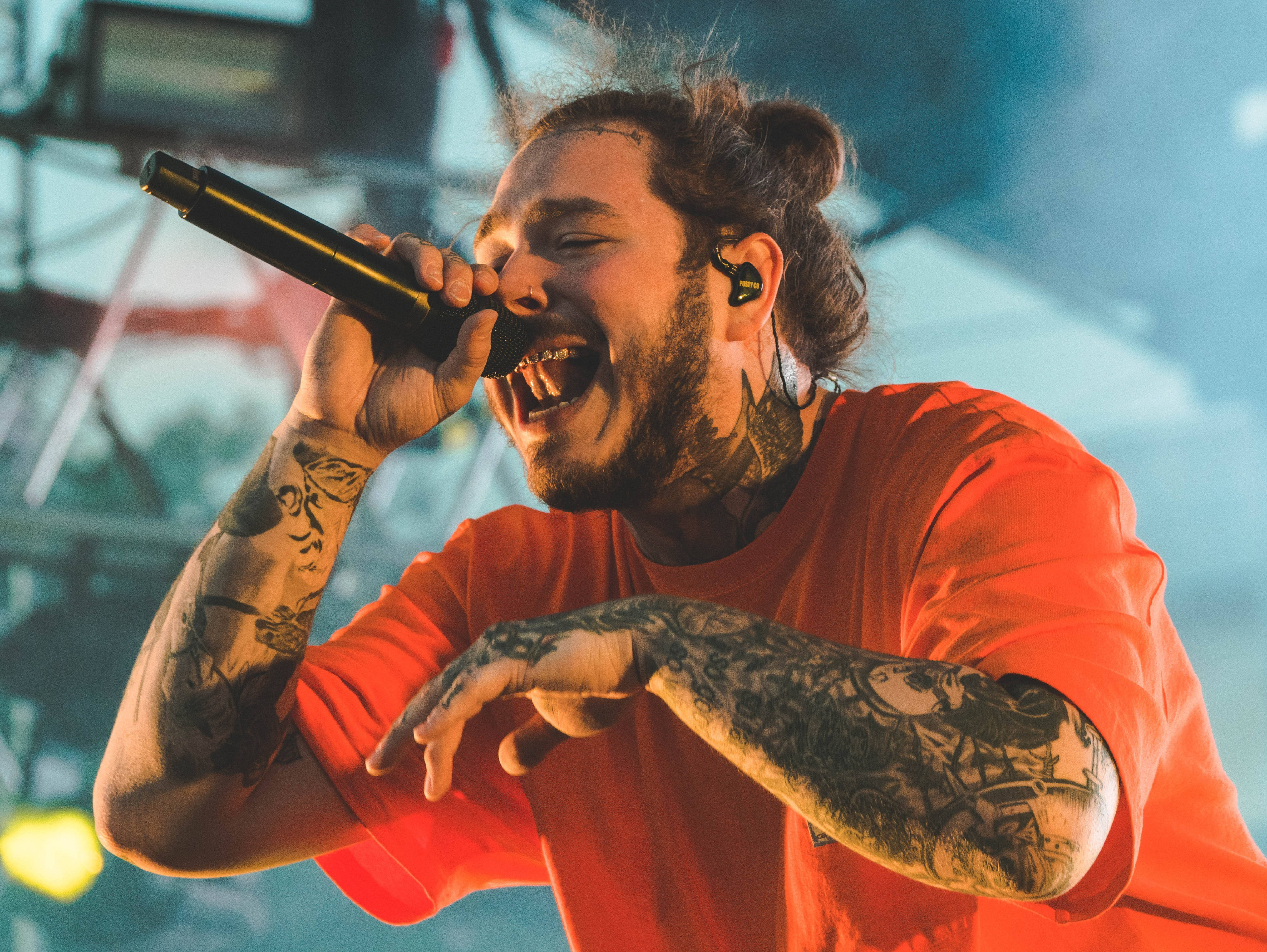 Post Malone 德克薩斯州 Posty Fest 壁紙 — 一位紋身男歌手身穿紅色襯衫，在燈光璀璨的舞台上熱情地對著麥克風演唱。