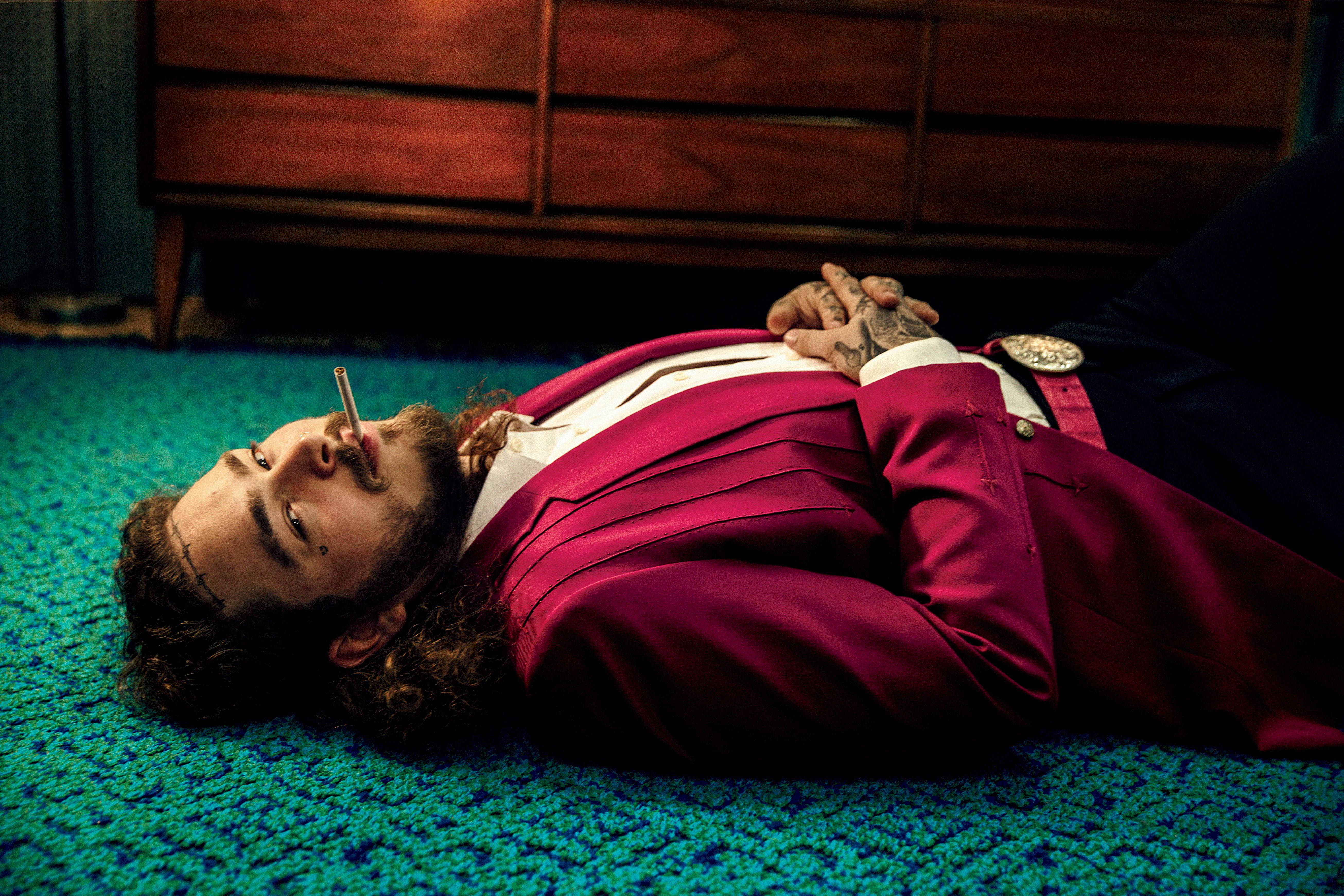 Post Malone《滾石》2017年11月封面 壁紙 — 一名身穿鮮豔洋紅色西裝的男子躺在藍綠色地毯上，嘴裡叼著香煙，紋身的手交叉放在胸前。