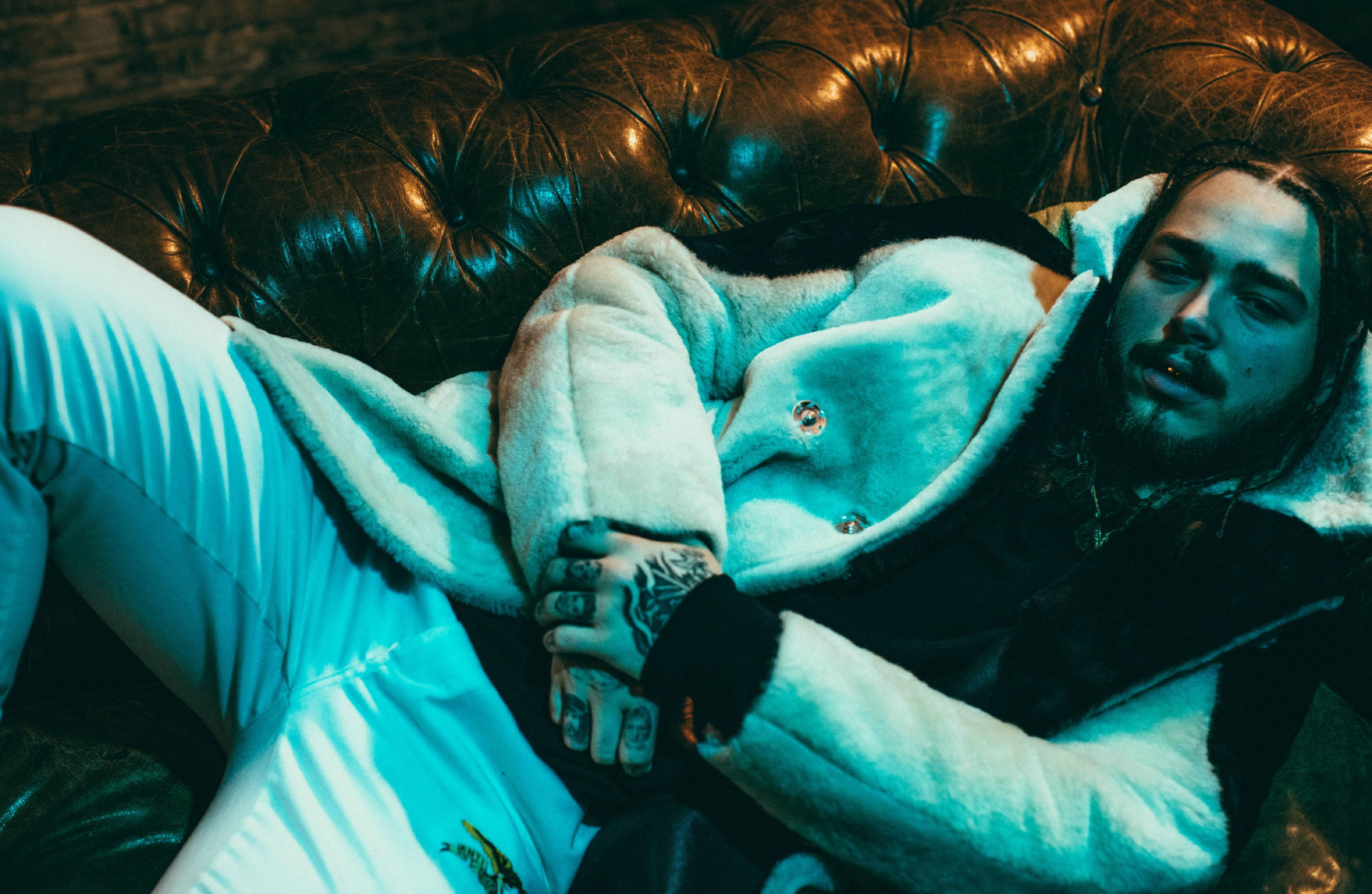 Post Malone《福布斯》2017年封面 壁紙 — 一個有紋身的男人在冷藍燈光下，穿著毛茸茸的白色外套，靠在一張簇絨皮革沙發上休息。