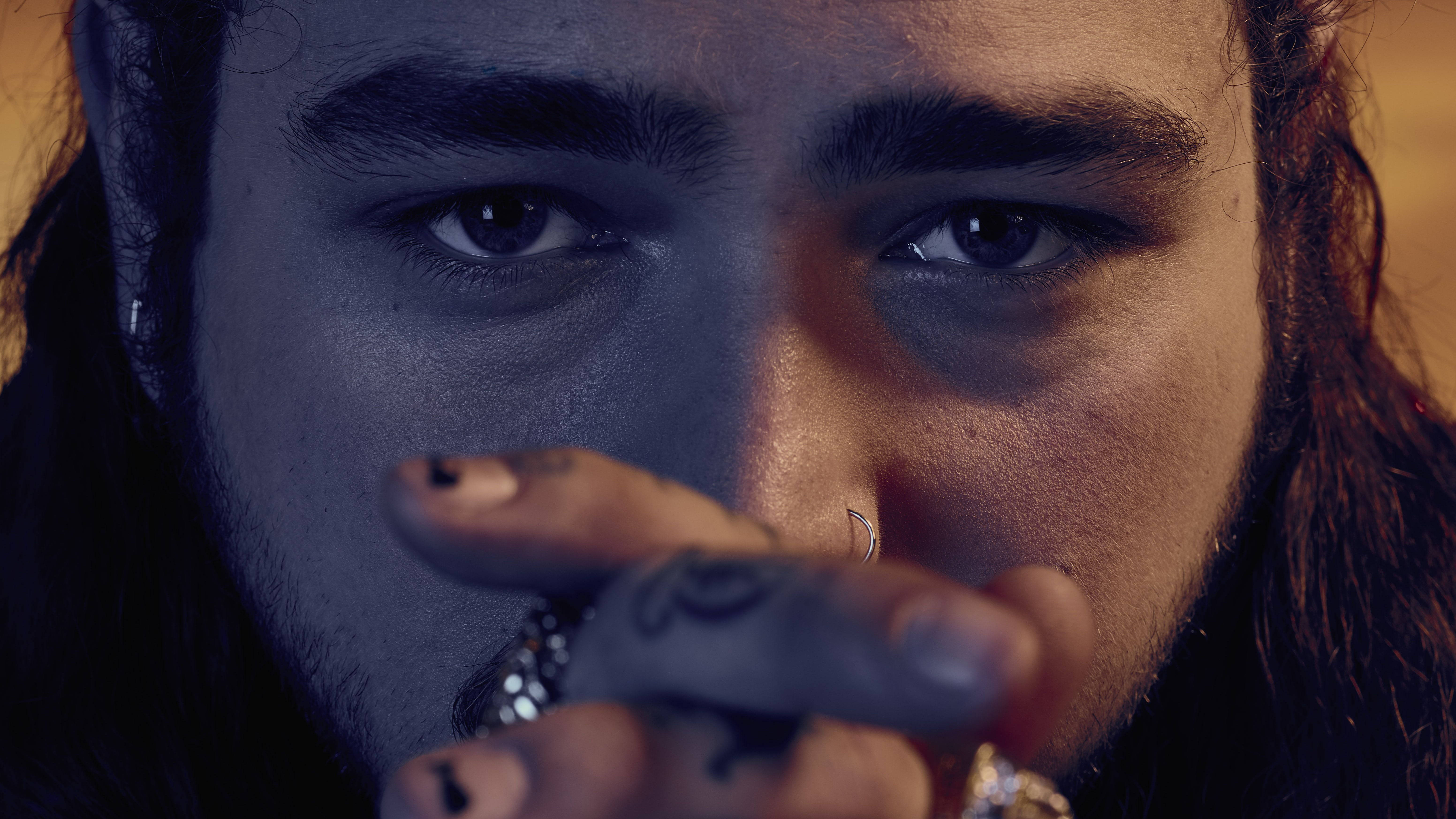 Post Malone 近照 壁紙 — 近距離肖像，一名眼神銳利、有紋身的男子，在戲劇性的藍色和橙色燈光下，將手放在臉旁。