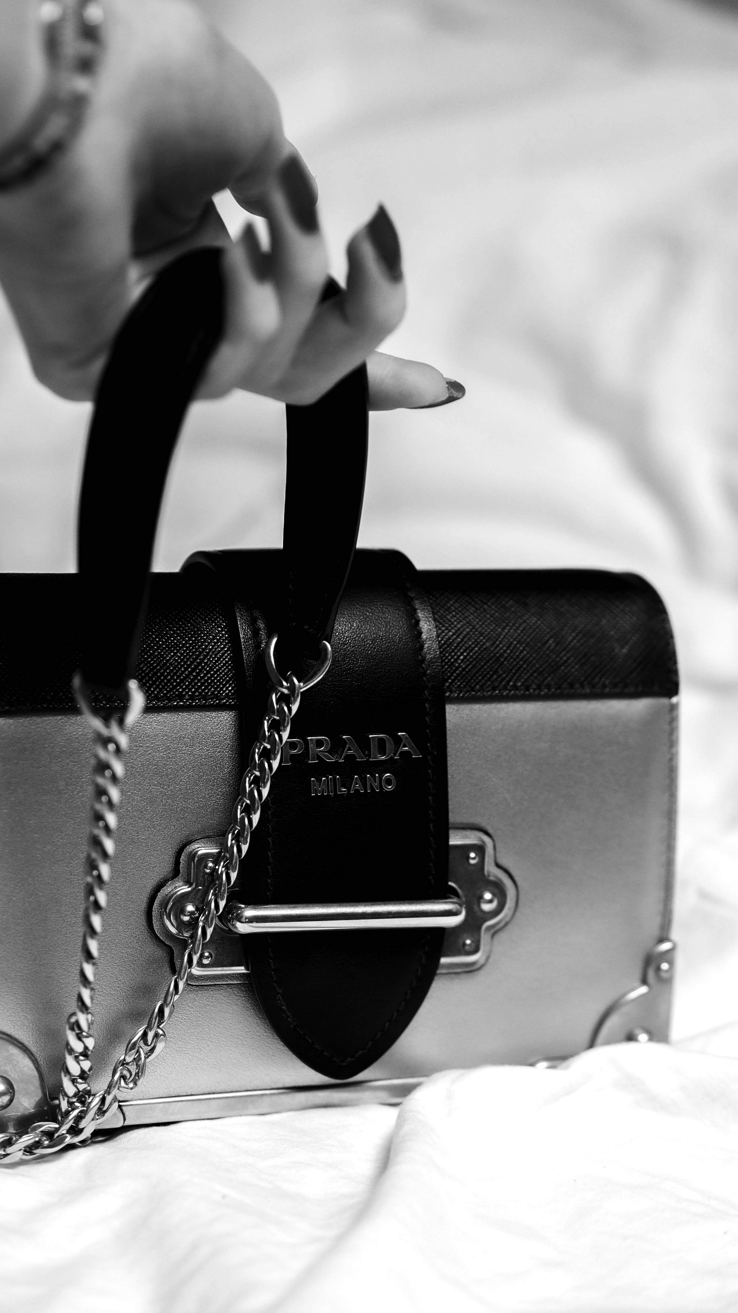 Prada 袋與尖指甲 壁紙 — 一隻塗有深色指甲油的手，在柔軟的白色布料背景上，以單色調拿著一個黑色和銀色的Prada手袋。