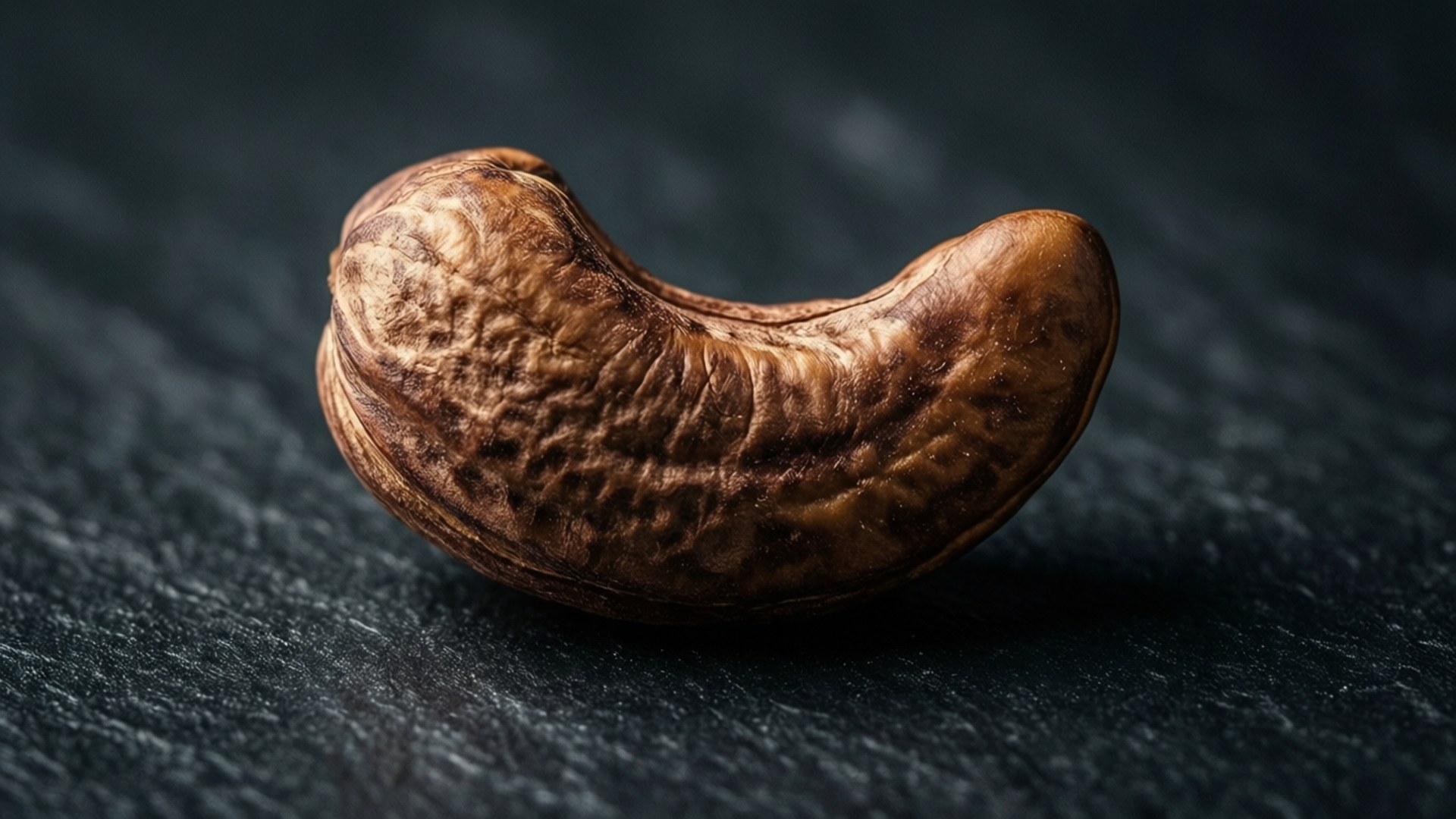 Premium Cashew Macro 壁紙 — 一顆帶有紋理的棕色腰果放在深色石板表面上，柔和的光線凸顯其彎曲形狀。