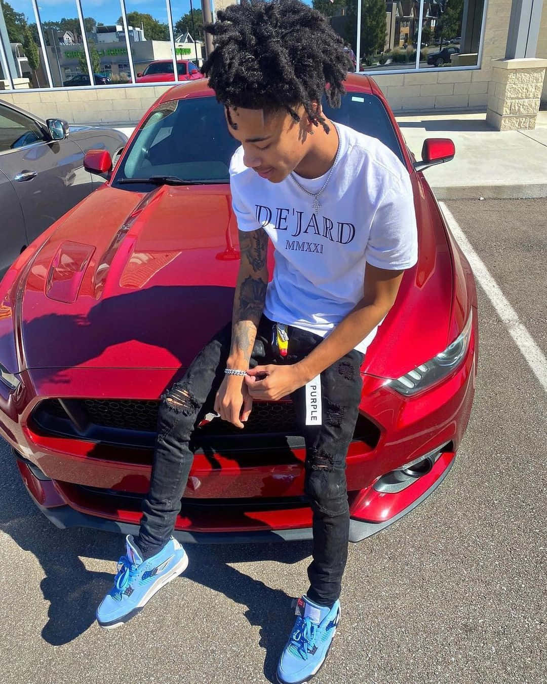 Prettyboyfredo 坐在紅色汽車上 壁紙 — 一個年輕男子留著捲髮，隨意地坐在陽光普照的停車場中一輛閃亮紅色跑車的引擎蓋上。