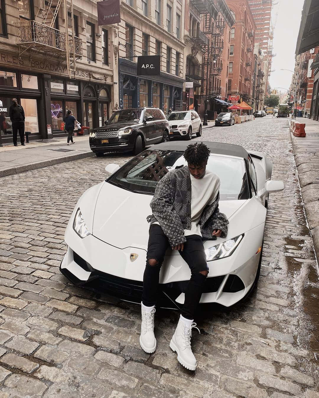 Prettyboyfredo 坐在白色蘭博基尼車上 壁紙 — 一名穿著白色靴子的年輕男子，在濕漉漉的鵝卵石街道上，靠著一輛白色林寶堅尼，街道兩旁是磚砌建築。