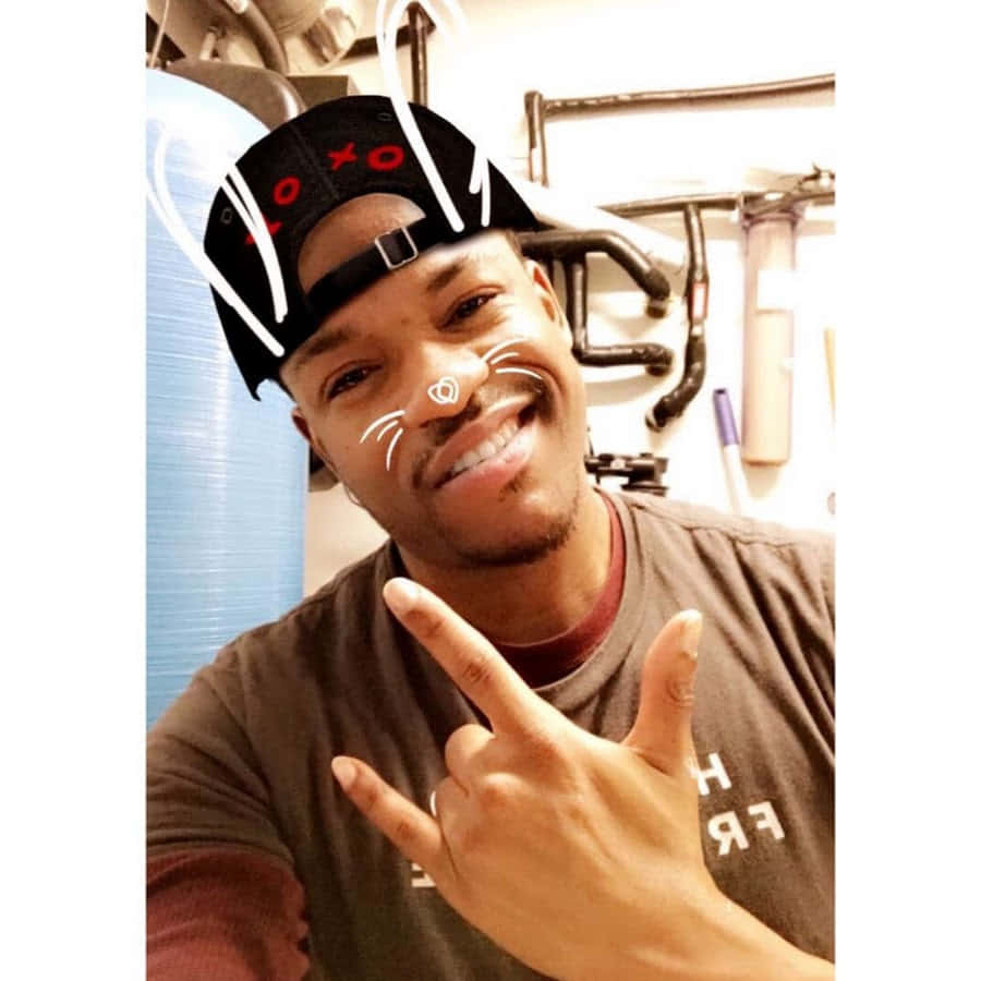 PrettyboyFredo 海灘休閒日 壁紙 — 一個微笑的男人戴著黑色帽子，上面有紅色XO文字和白色貓鬚濾鏡，在室內做出搖滾手勢。