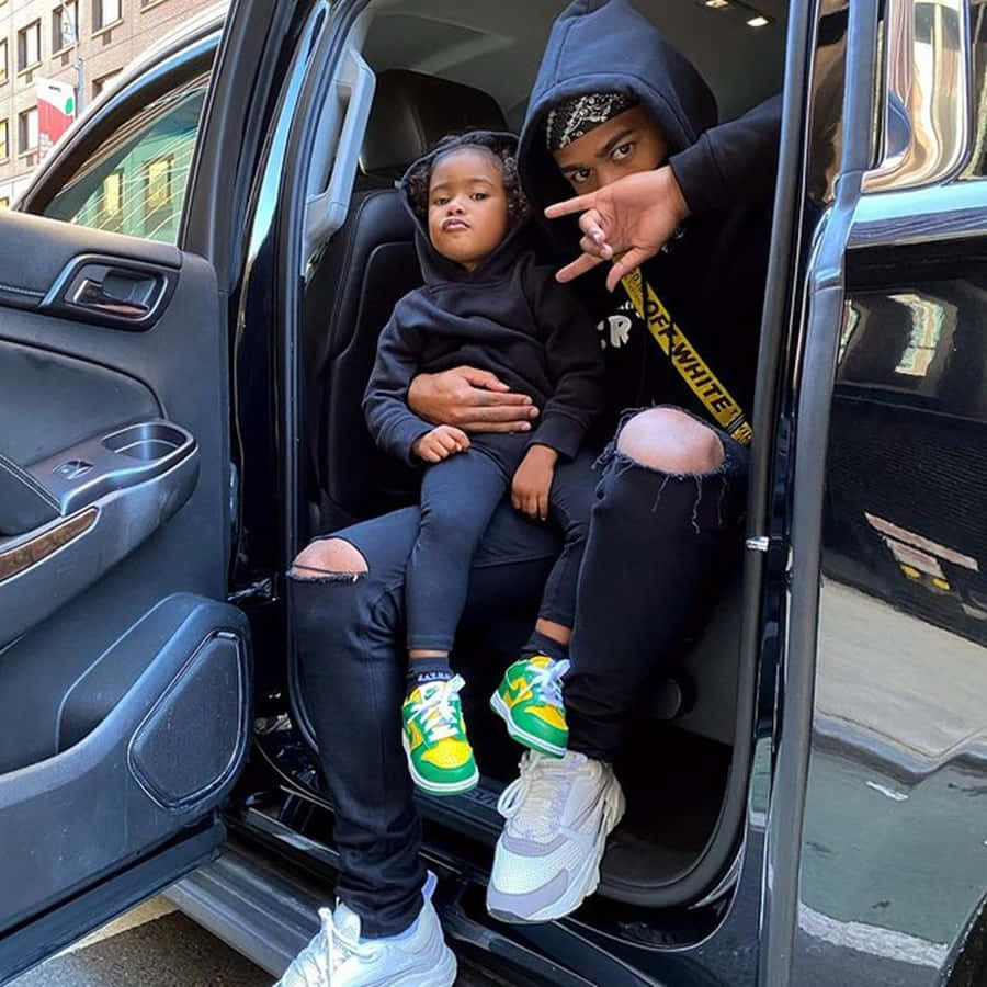 Prettyboyfredo 與女兒在車內 壁紙 — 一個大人和一個小孩穿著黑色連帽衫坐在車門邊，擺出各種手勢，腳穿色彩繽紛的運動鞋。