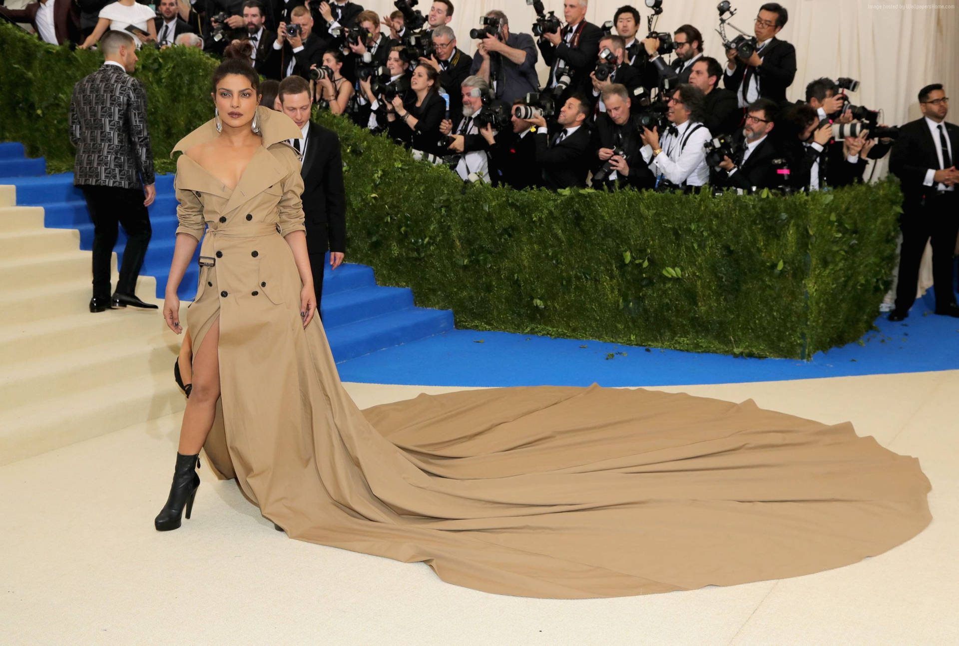 Priyanka Chopra 在一個晚會上穿著米色風衣長裙站在藍色地毯上。