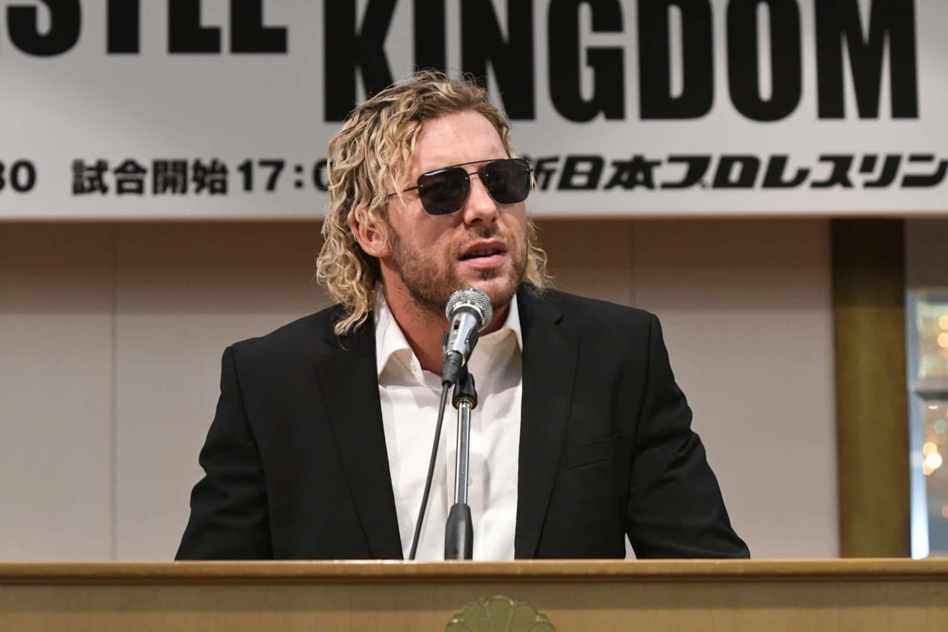 身穿西裝、戴著太陽眼鏡的男子在帶有「KINGDOM」橫幅的記者會上發言。