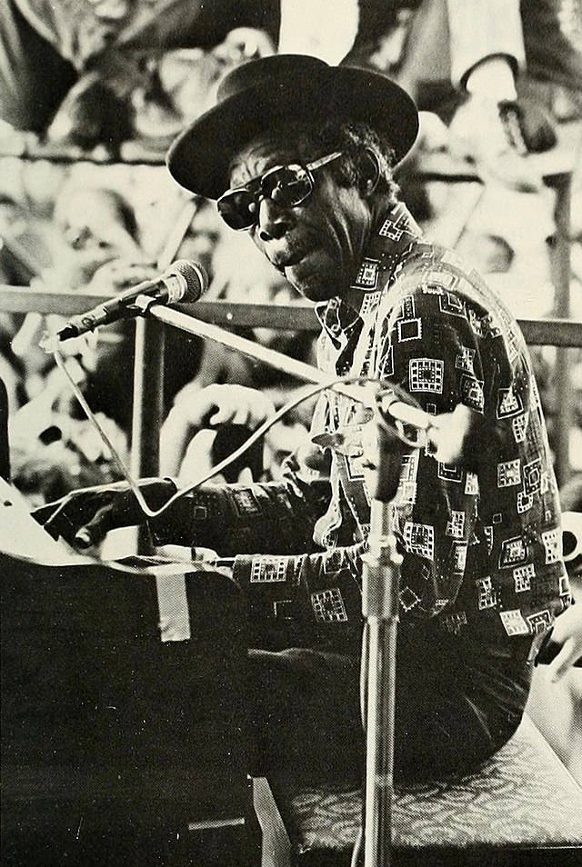 Professor Longhair 1975年新奧爾良爵士與遺產節 壁紙 — 一張黑白照片，一位戴帽子和太陽眼鏡的爵士鋼琴家正在用麥克風彈鋼琴。