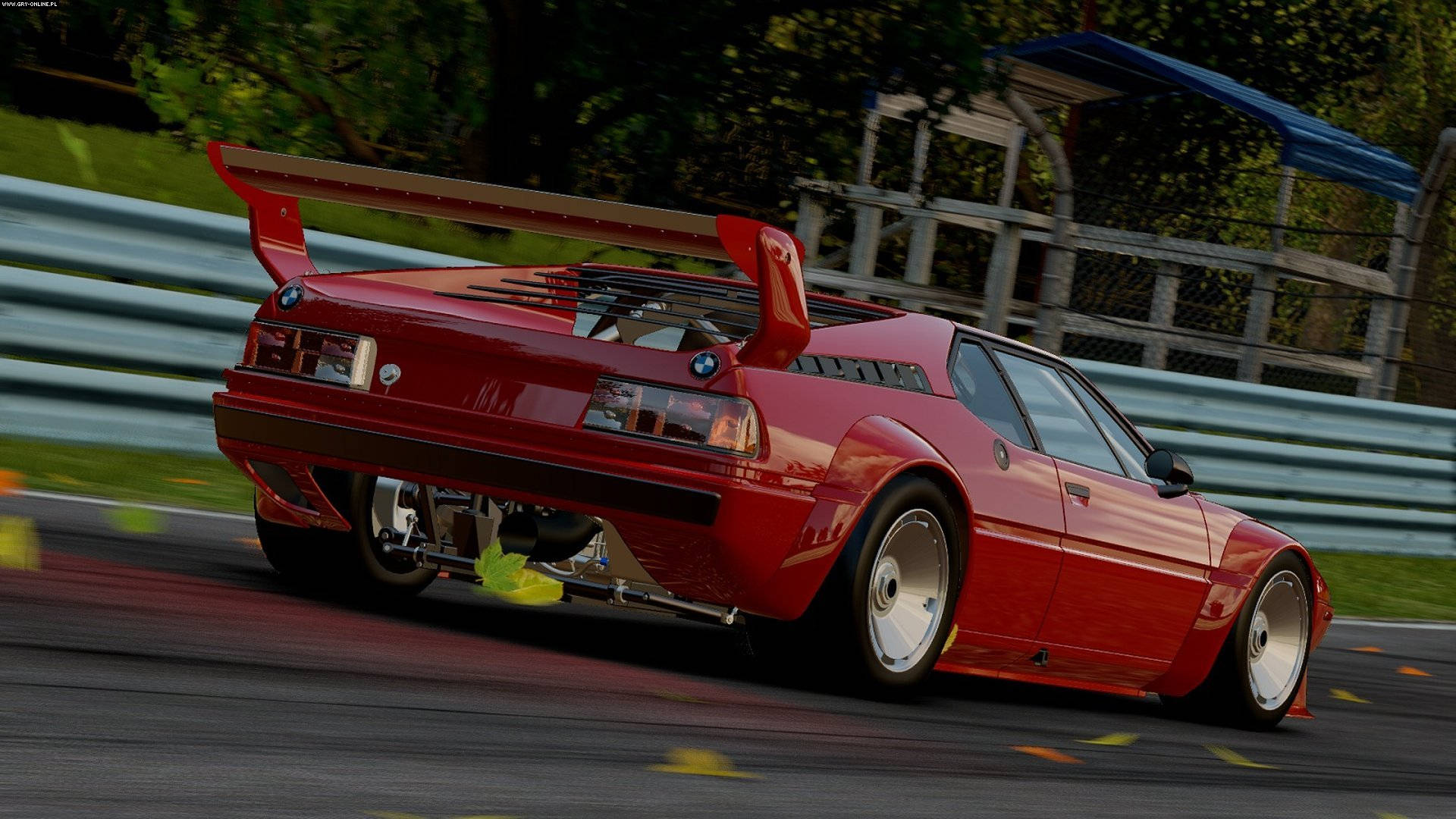 Project Cars 4K 紅色寶馬 壁紙 — 一輛鮮紅色、帶有大型尾翼的BMW M1賽車，在綠樹和護欄環繞的賽道上疾馳。