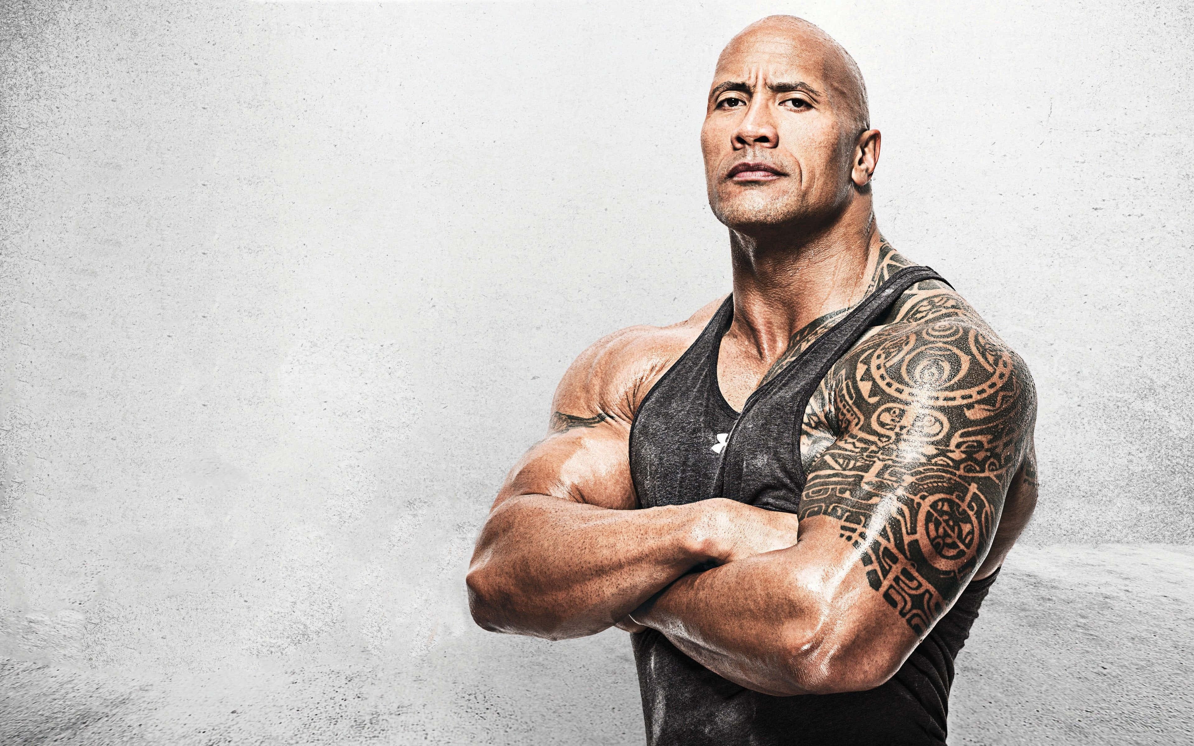 黑色 Project Rock Sando 壁紙 — 一個有紋身的肌肉男自信地交叉雙臂，靠著紋理灰色牆壁站立。