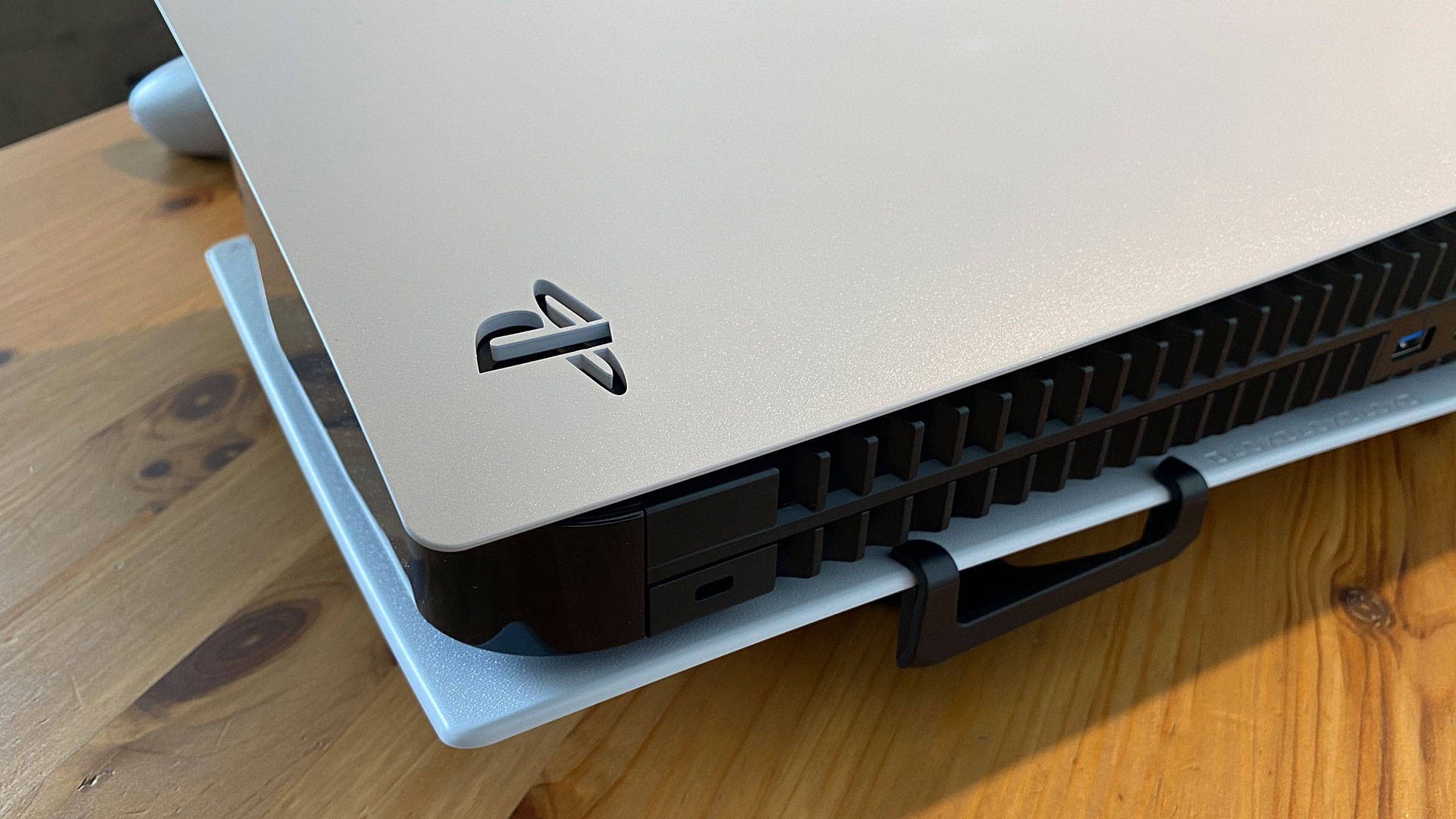 PS5盒裝主機 壁紙 — 銀色PlayStation 5主機特寫，放置於淺藍色底座上，底座置於溫暖的木桌上。