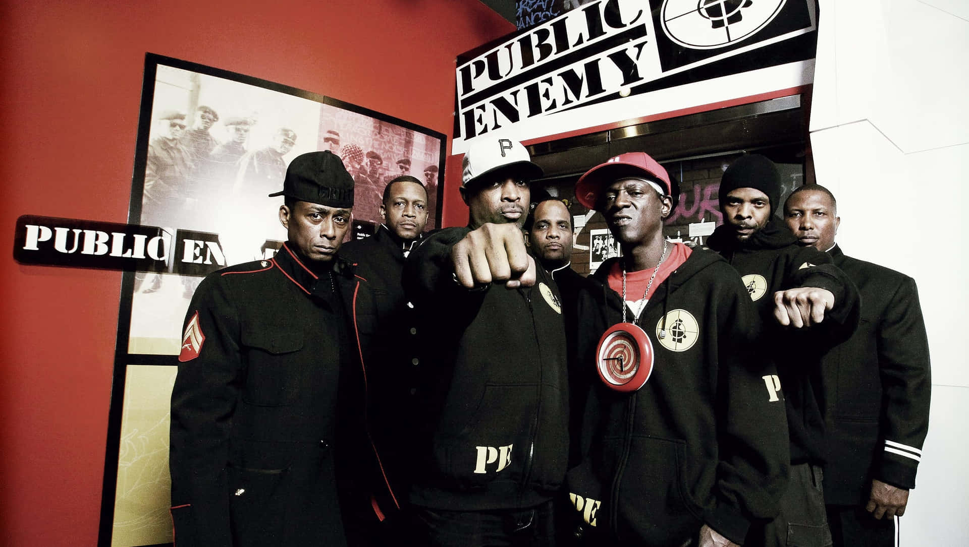  壁紙 — Public Enemy 樂隊成員身穿帶有紅色點綴的黑色外套，在他們標誌性的標誌和裱框照片前挑釁地擺姿勢。