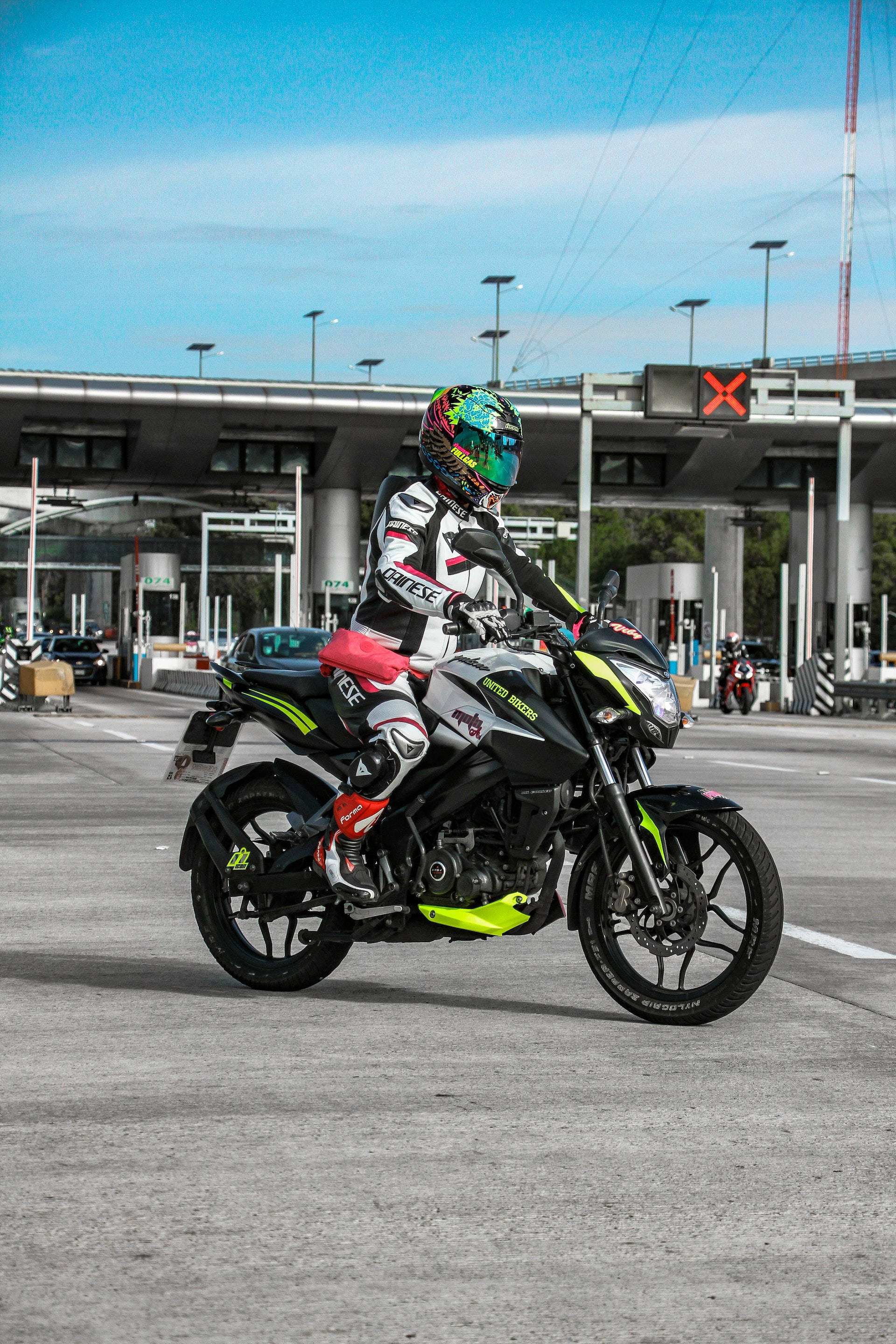 霓虹Pulsar RS200 壁紙 — 一名全副裝備的電單車手在晴朗的藍天下，於高速公路收費站騎著一輛運動型黑色和螢光綠色的電單車。