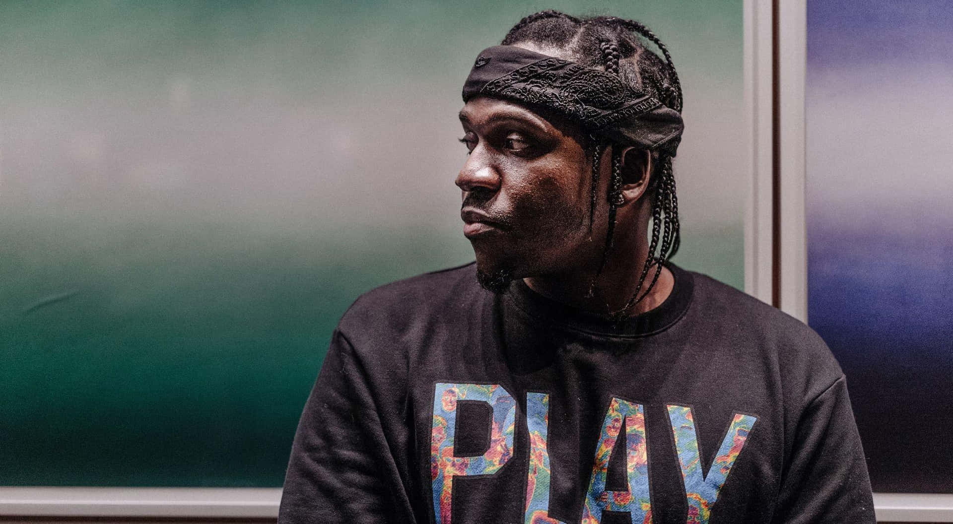 Pusha T戴黑頭帶穿Play字樣上衣 壁紙 — 一名辮子頭髮、戴頭帶的男子，穿著帶有彩色「PLAY」圖案的黑色運動衫，若有所思地凝視著。