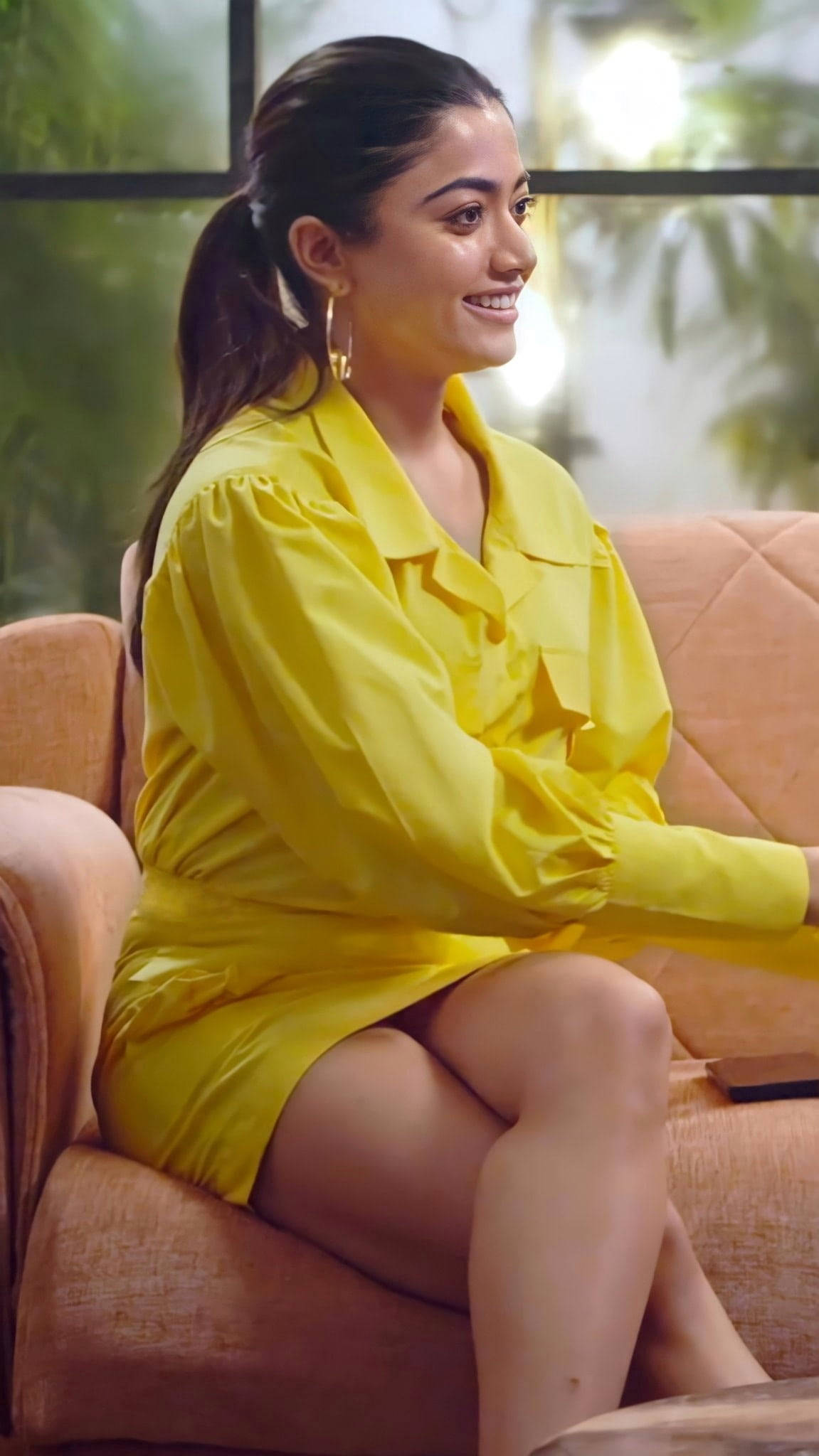 《Pushpa》女主角拉什米卡·曼丹納的優雅造型 壁紙 — 一位身穿亮黃色連衣裙的女性坐在米色沙發上微笑，身後窗戶可見綠色植物。