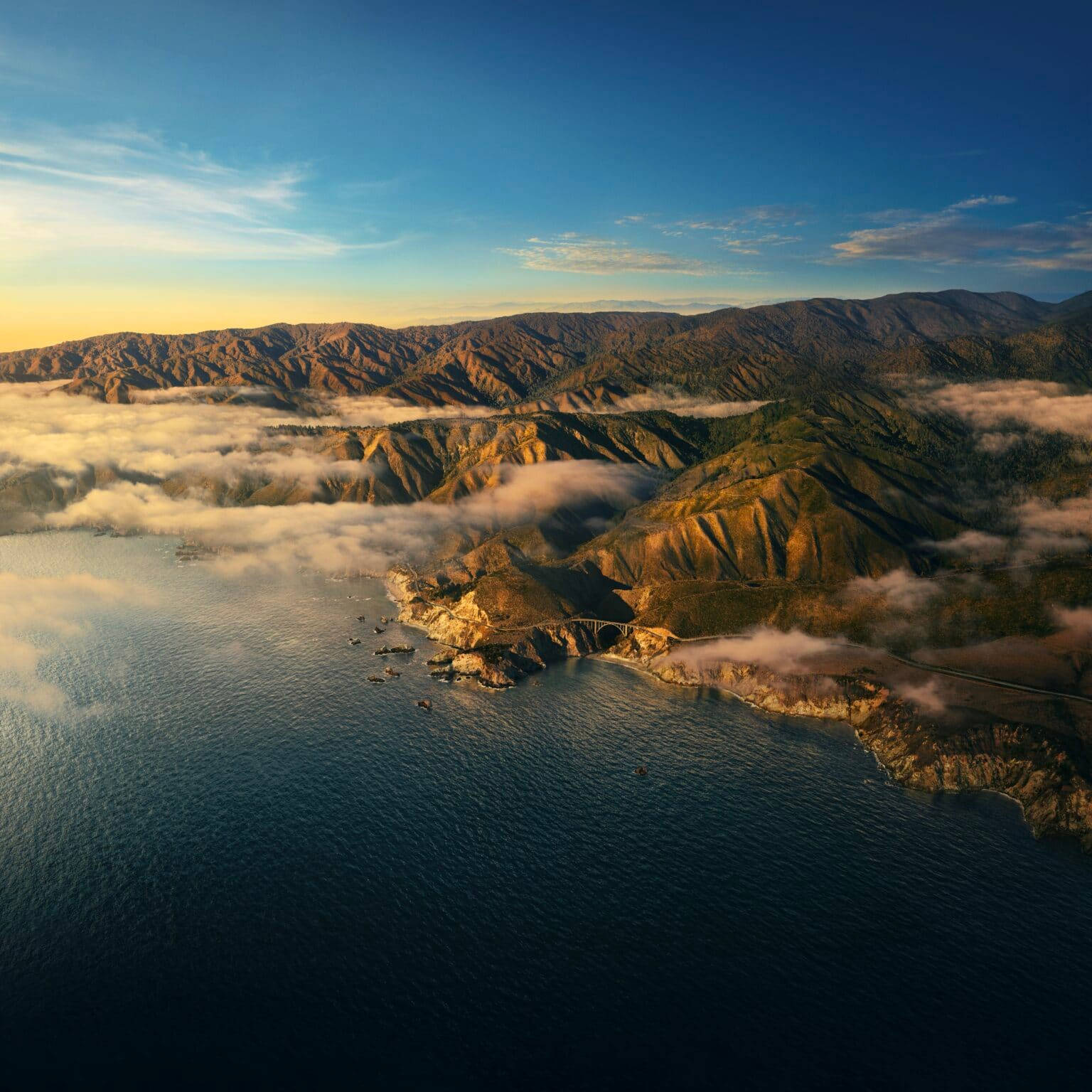 Big Sur空中壯麗景觀 壁紙 — 崎嶇海岸山脈的鳥瞰圖，沐浴在金色日出光芒中，雲霧繚繞山谷，下方是深藍色海洋。