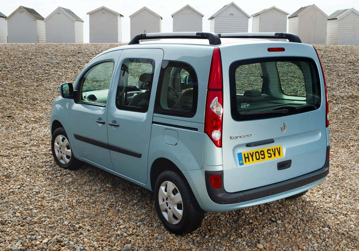 Renault Kangoo 桌布 壁紙 — 一輛淺藍色的雷諾Kangoo客貨車停泊在卵石灘上，背景有白色沙灘小屋。