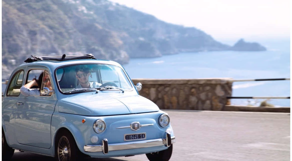 Fiat 500 桌布 壁紙 — 一輛淺藍色復古菲亞特500沿著海岸懸崖道路行駛，一名男子駕駛，一名女子從乘客座位揮手。