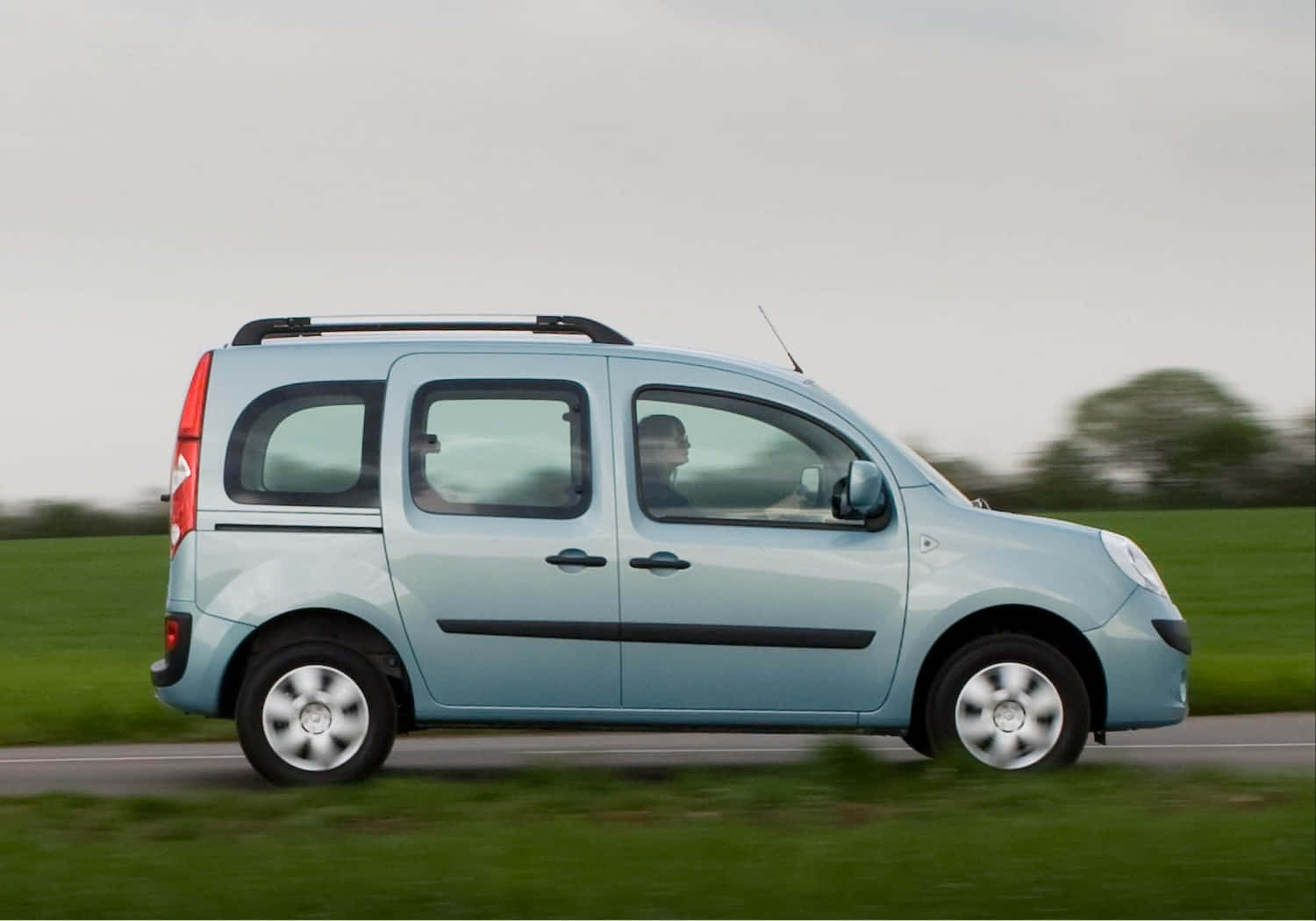 Renault Kangoo 桌布 壁紙 — 一輛淺藍色緊湊型貨車配備車頂行李架，在模糊的綠色田野和多雲天空的鄉村道路上行駛。