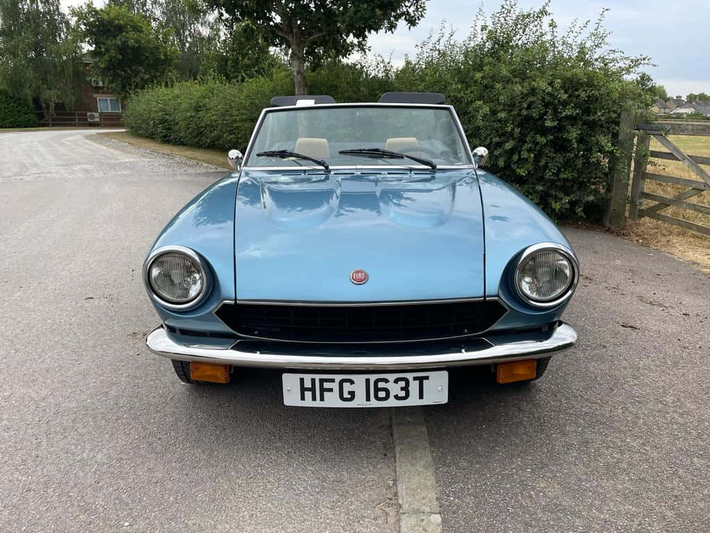 Fiat 124 Spider 桌布 壁紙 — 一架淺藍色經典菲亞特124 Spider開篷車停泊在碎石車道上，背景有綠色籬笆和木門。