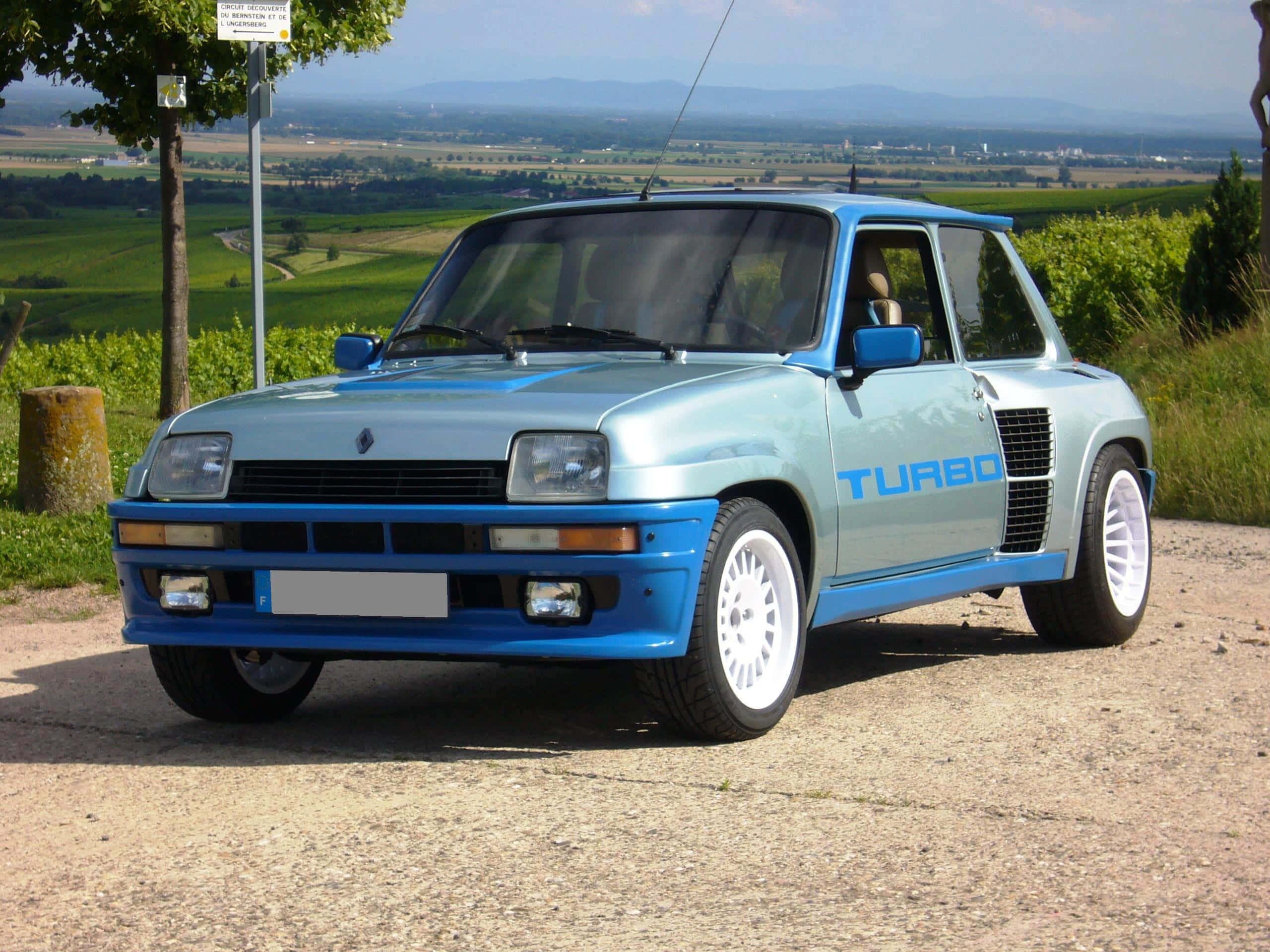 Renault 5 Turbo 桌布 壁紙 — 一輛淺藍色雷諾5 Turbo配白色輪胎，停泊在碎石路上，俯瞰著晴朗天空下的綠色起伏山丘和遠山。