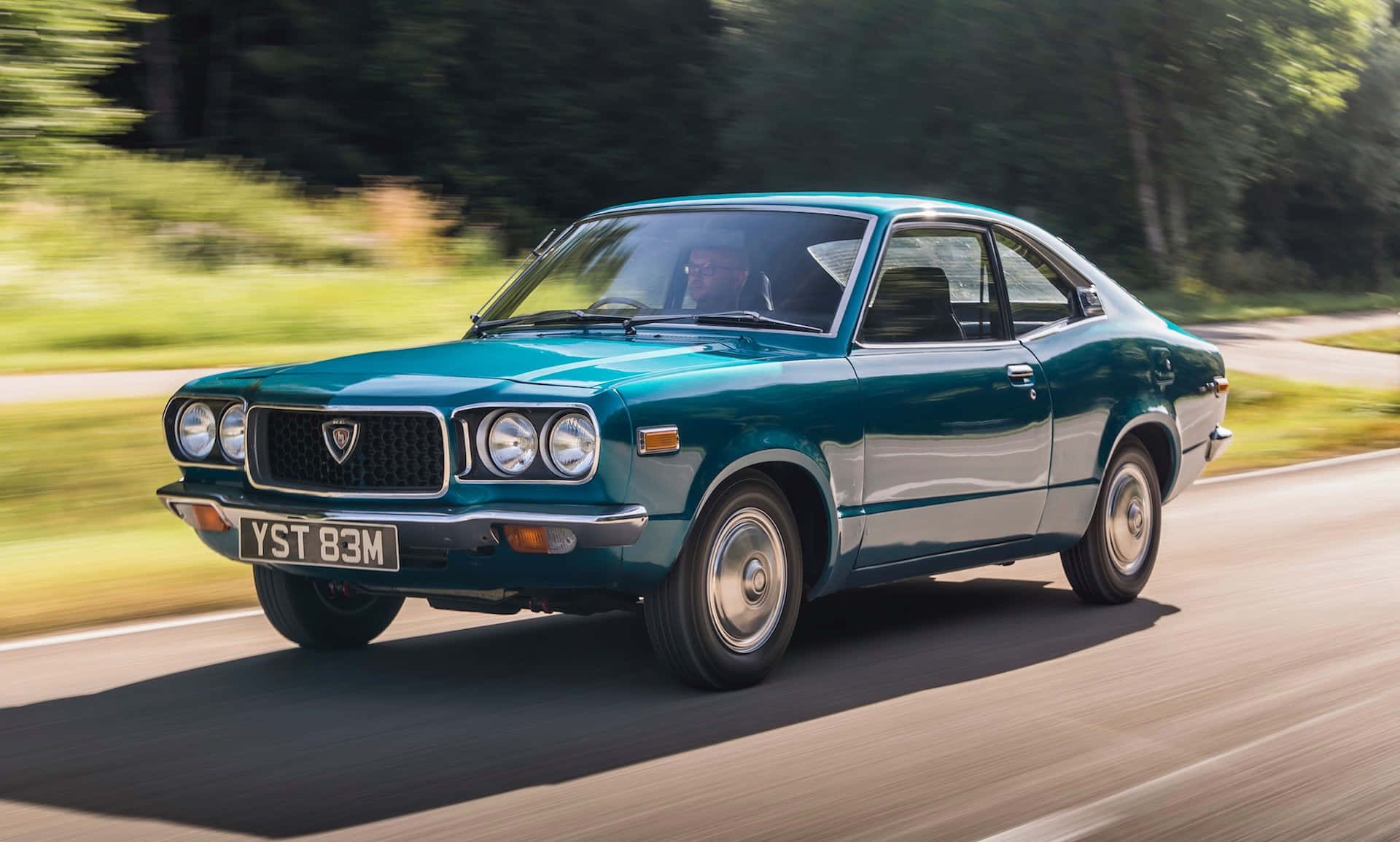 Mazda Rx-3 桌布 壁紙 — 一輛青綠色經典汽車在陽光明媚的道路上行駛，背景有模糊的綠色樹木。