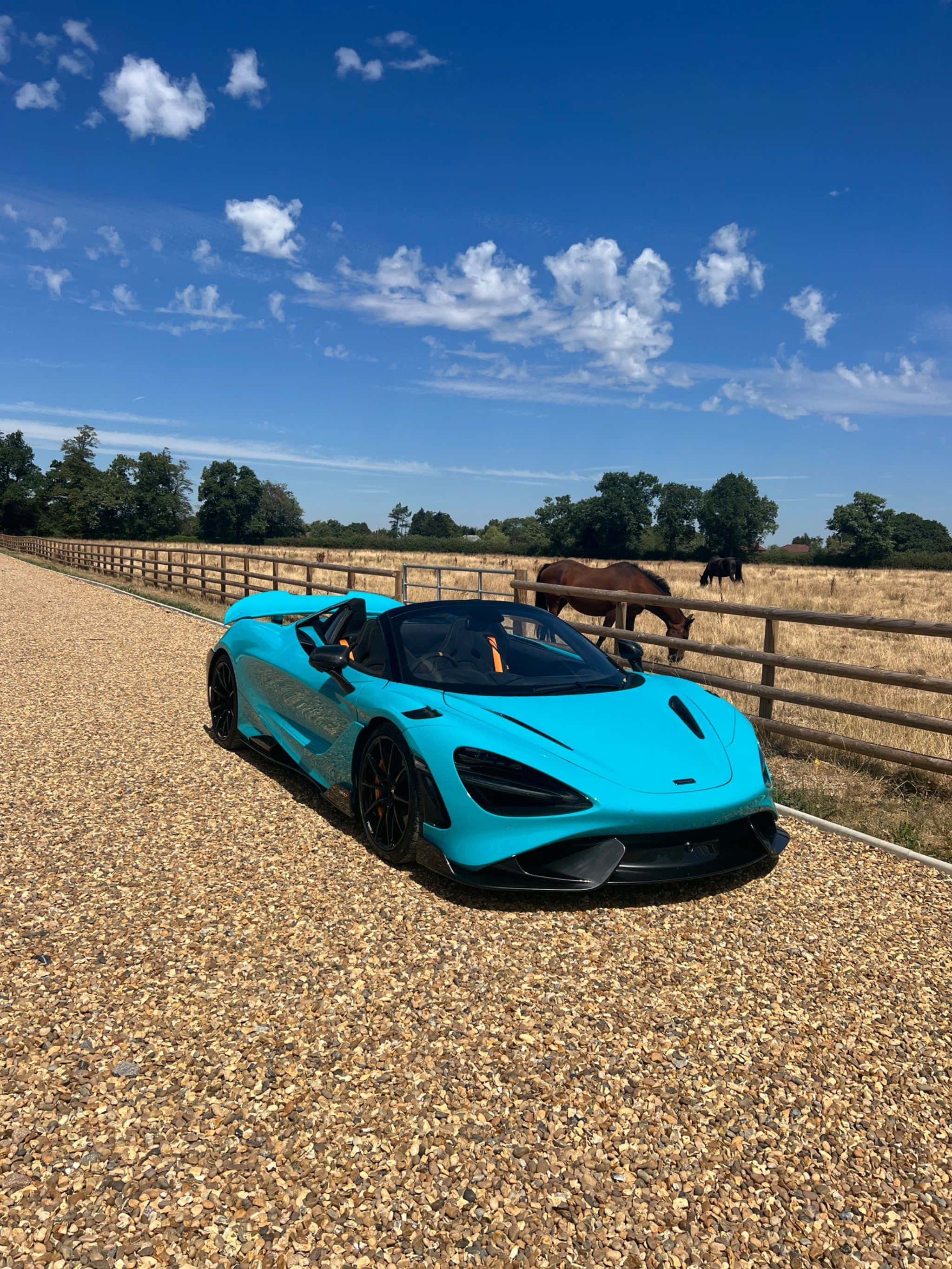 Mclaren 765lt 桌布 壁紙 — 一輛充滿活力的青綠色McLaren跑車停泊在碎石車道上，旁邊是圍欄牧場，馬匹在晴朗藍天和零星白雲下吃草。
