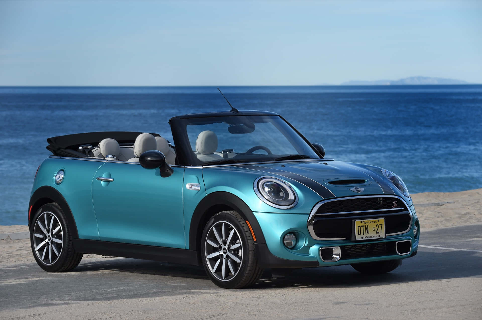 Mini Cooper Convertible 桌布 壁紙 — 一架青綠色MINI Cooper開篷車停泊在陽光明媚的海灘上，海灘上有海浪和遠處的山脈，天空清澈湛藍。