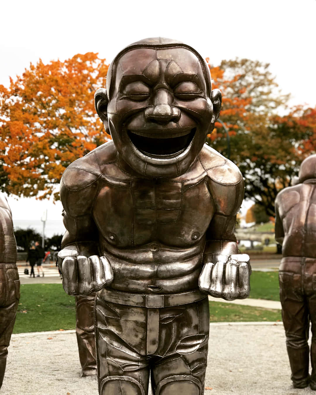  壁紙 — 一個肌肉發達、大笑的男人的大型銅像矗立在公園裡，後面有秋天的樹木。